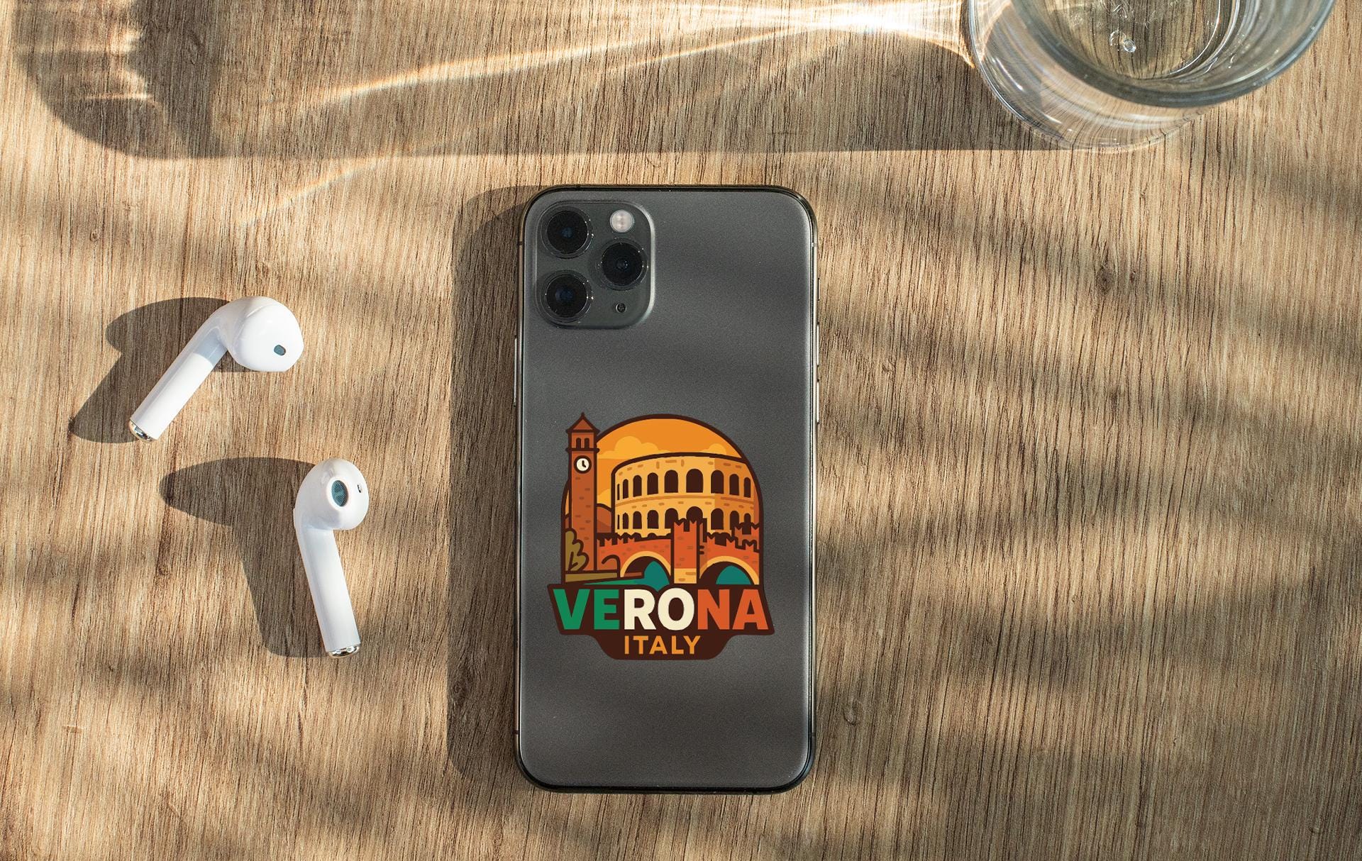 a smartphone with a sticker of the Colosseum on it, placed on a wooden surface alongside a pair of white earbuds and a glass of water.