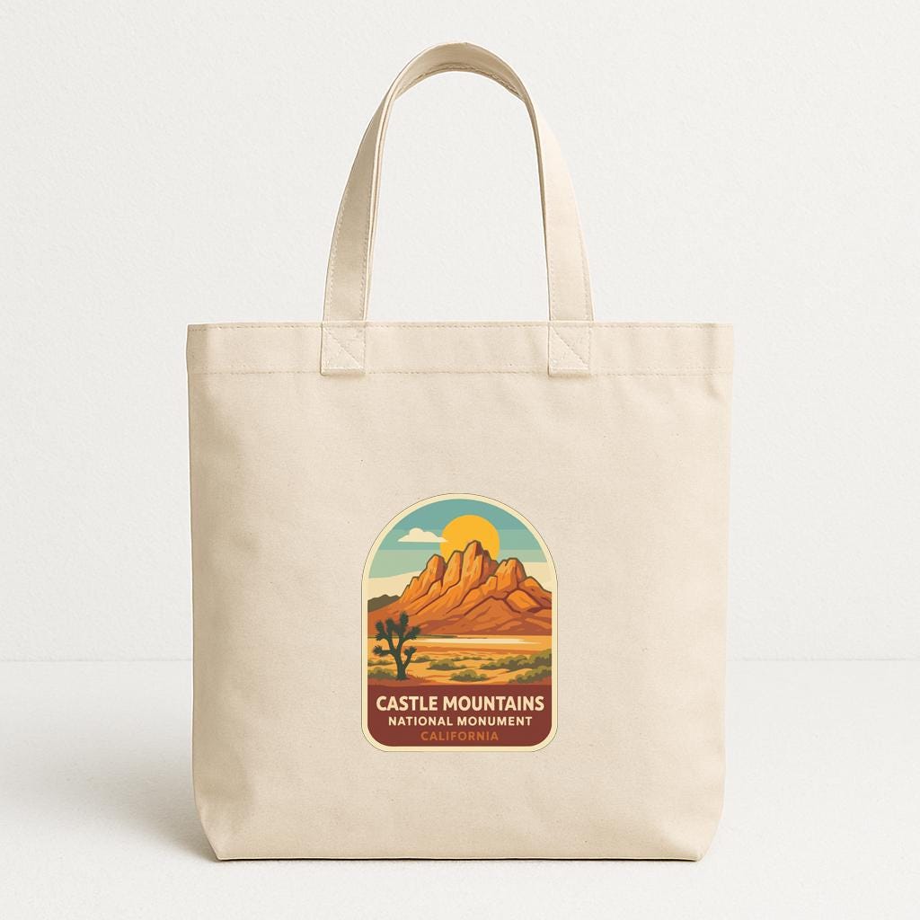 A beige tote bag with a graphic design featuring a desert landscape with mountains and a cactus, along with the text "CASTLE MOUNTAINS NATIONAL MONUMENT CALIFORNIA".