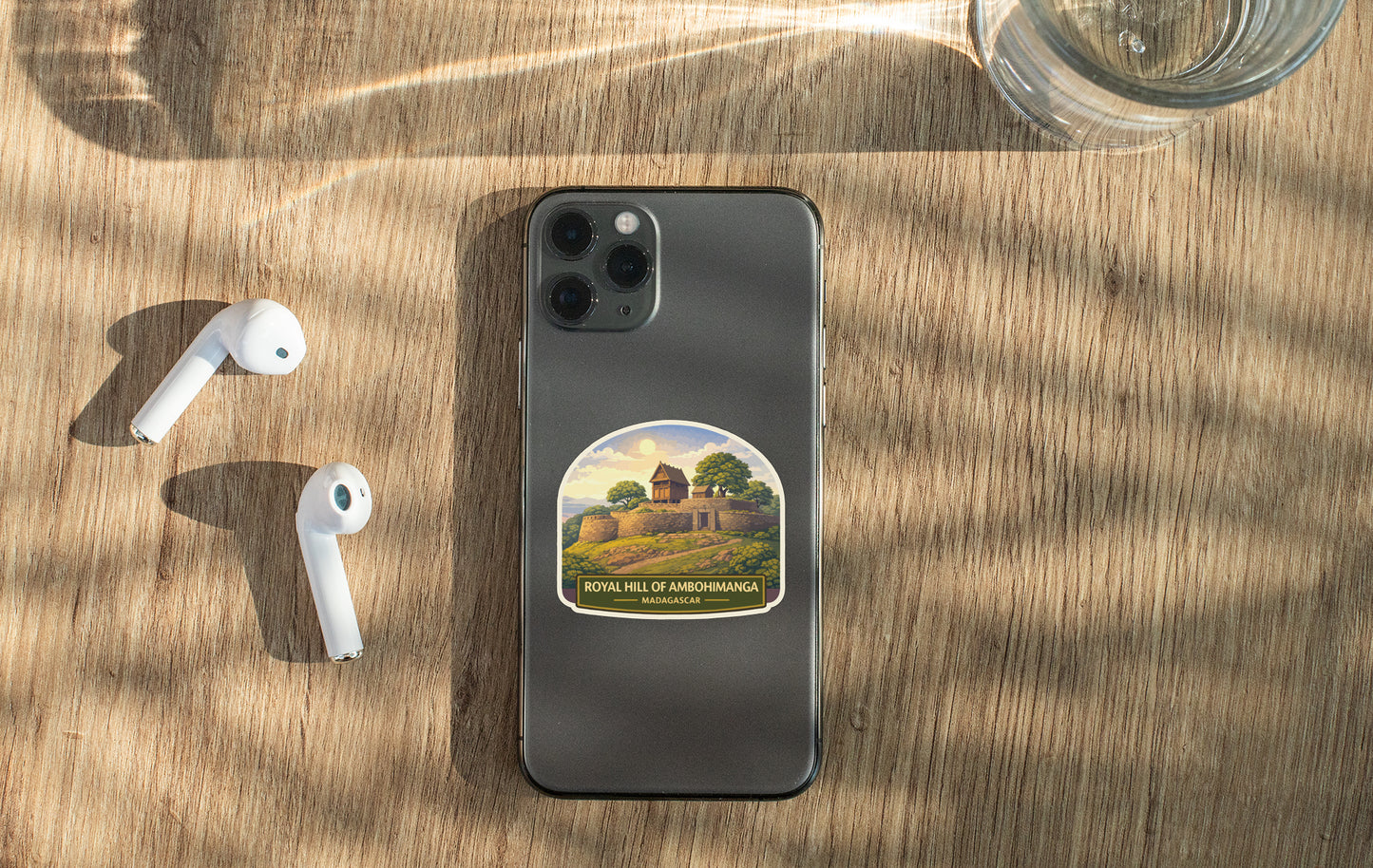 a smartphone with a sticker on the back, a pair of white earbuds, and a glass of water on a wooden surface.