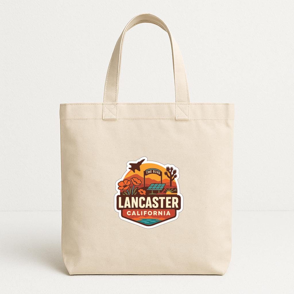 A beige tote bag with a graphic design featuring a sunset, palm trees, and the text "LANCASTER CALIFORNIA".