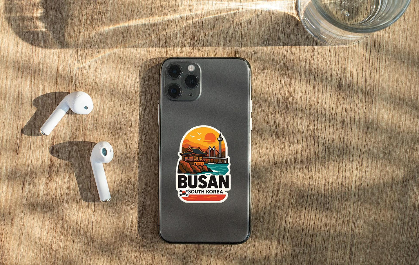 a smartphone with a sticker on the back, placed on a wooden surface alongside a pair of white earbuds and a glass of water.