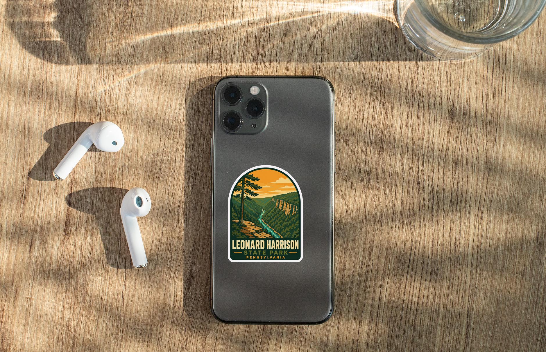 A smartphone with a sticker of a scenic landscape on the back, along with a pair of white earbuds and a glass of water on a wooden surface.