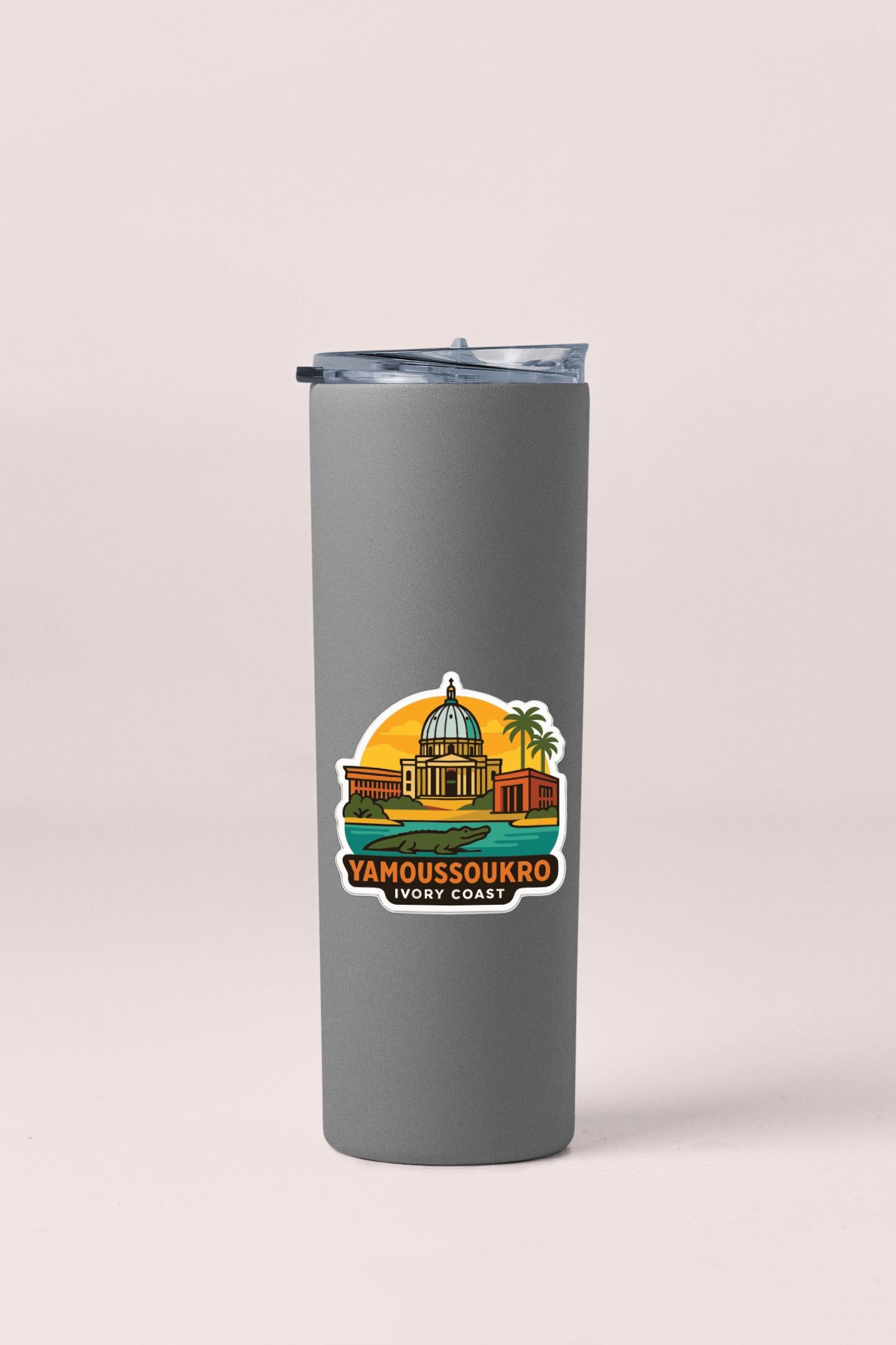 A gray insulated tumbler with a graphic design depicting a building and palm trees, along with the text "YAMOUSOUKRO IWYO COAST".