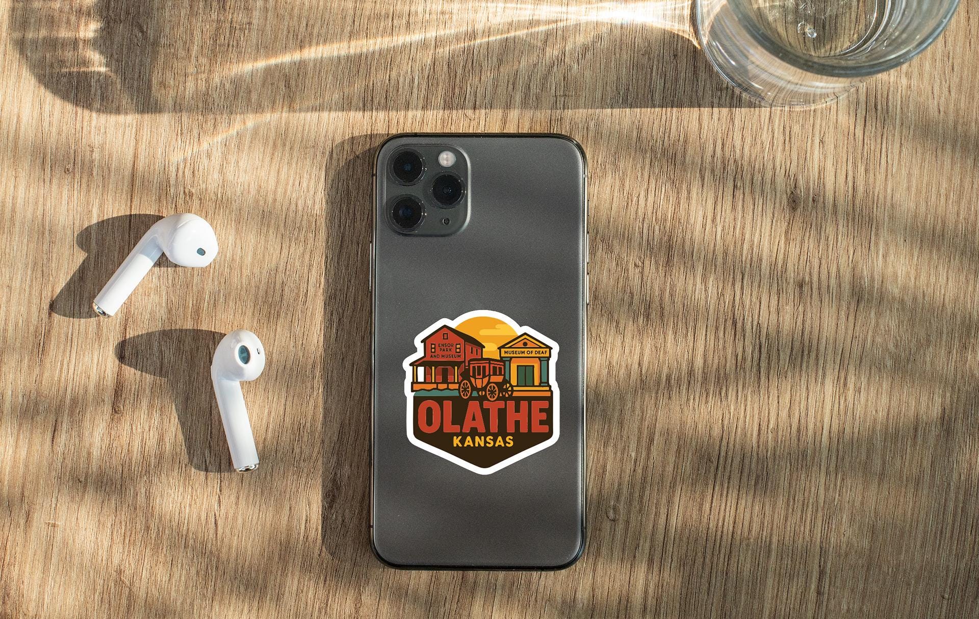a smartphone with a sticker on the back, placed on a wooden surface alongside a pair of white earbuds and a glass of water.