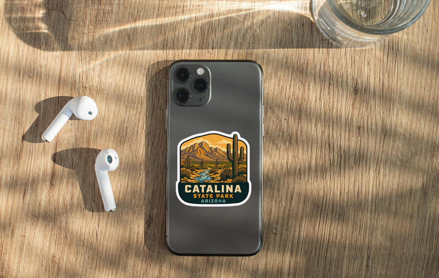 a smartphone with a sticker of Catalina Park, Arizona, placed on a wooden surface alongside a pair of white earbuds and a glass of water.
