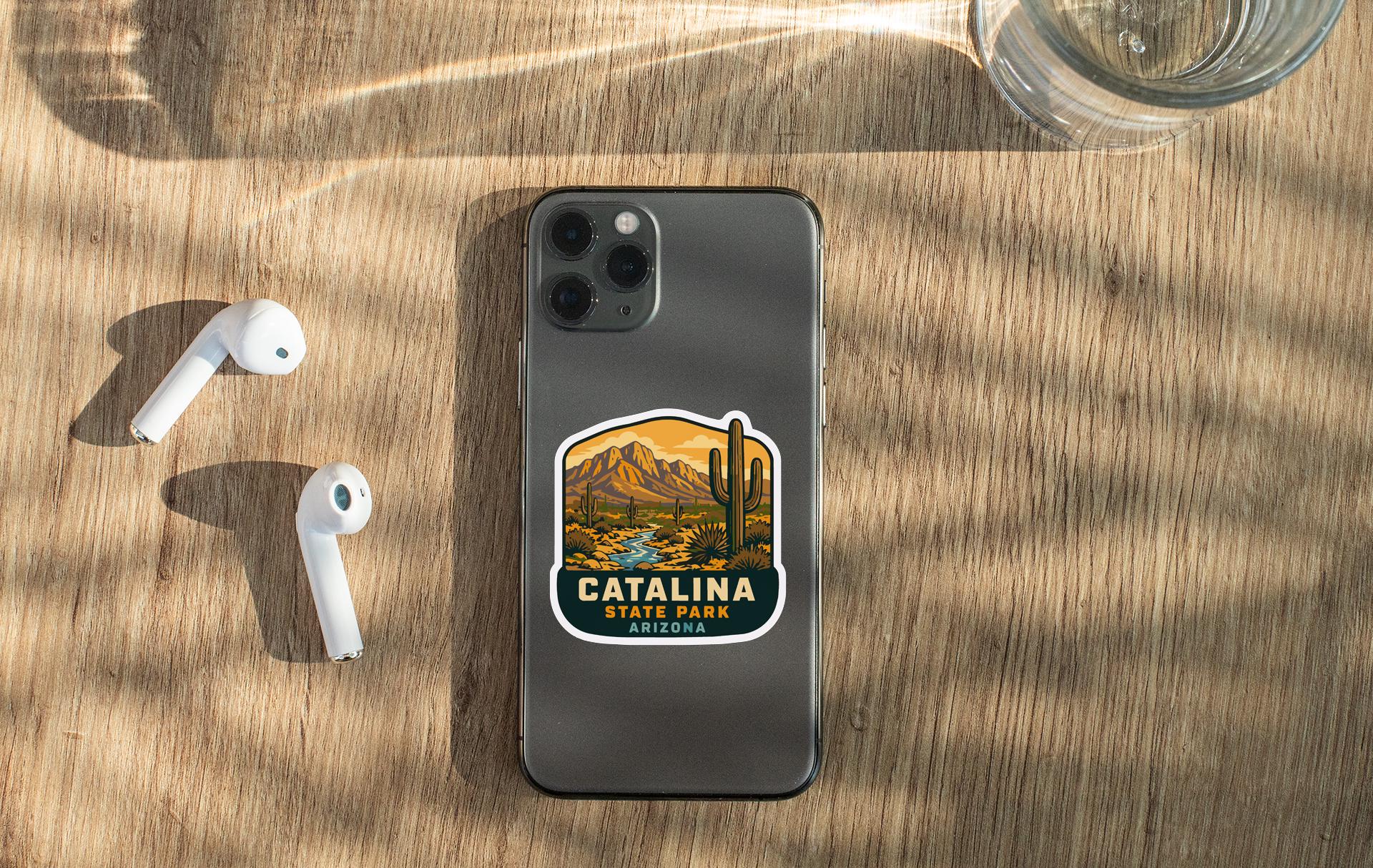 a smartphone with a sticker of Catalina Park, Arizona, placed on a wooden surface alongside a pair of white earbuds and a glass of water.