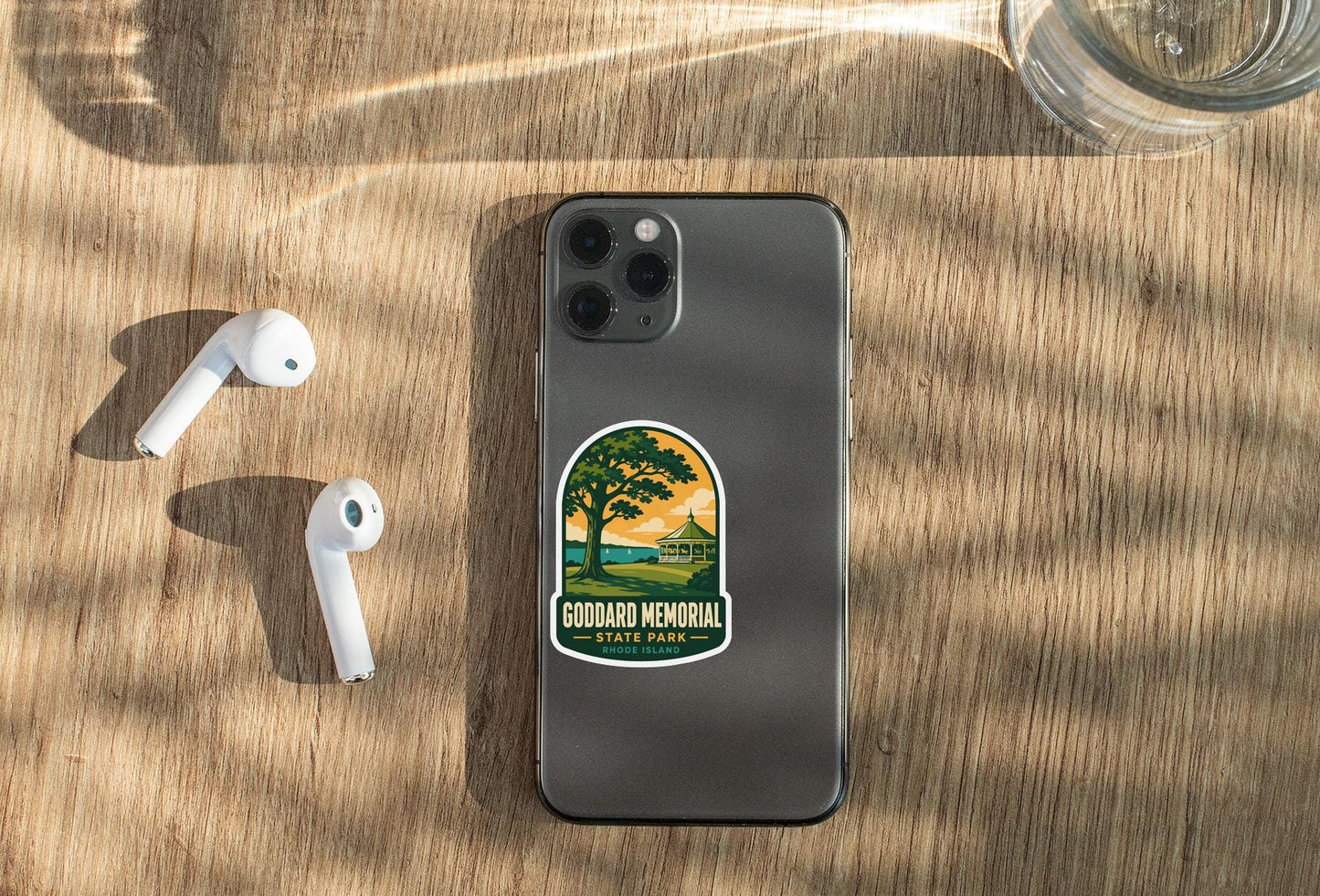 a smartphone with a sticker on it, placed on a wooden surface alongside a pair of white earbuds and a glass of water.