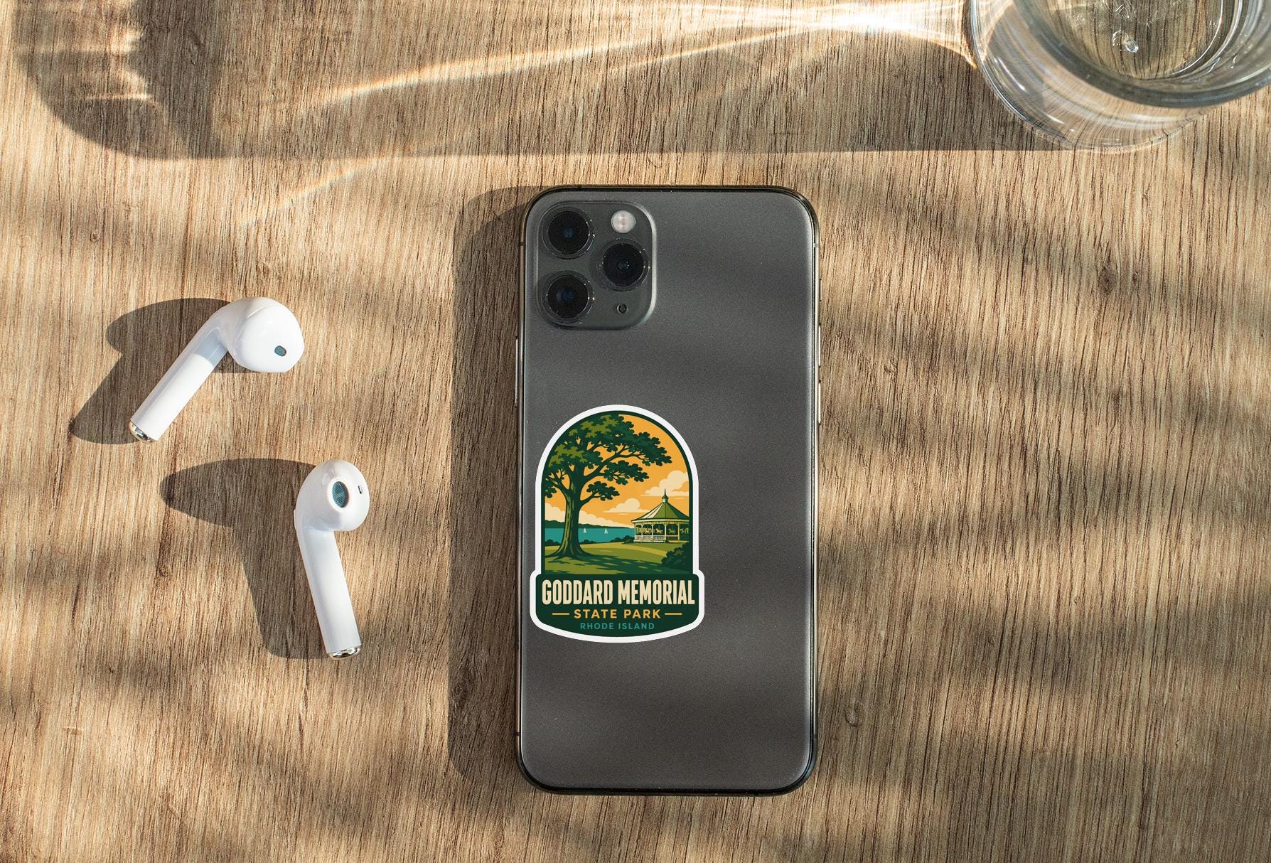 a smartphone with a sticker on it, placed on a wooden surface alongside a pair of white earbuds and a glass of water.