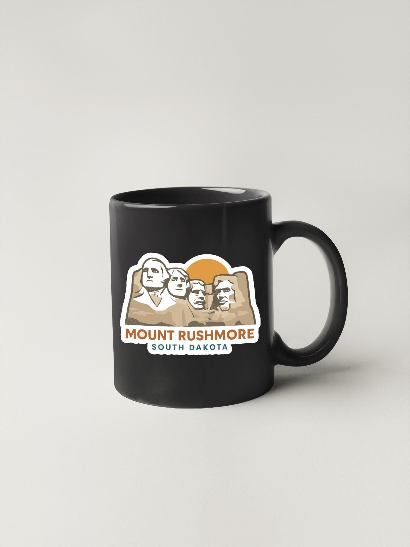 A black coffee mug with a graphic design featuring the Mount Rushmore monument in South Dakota.