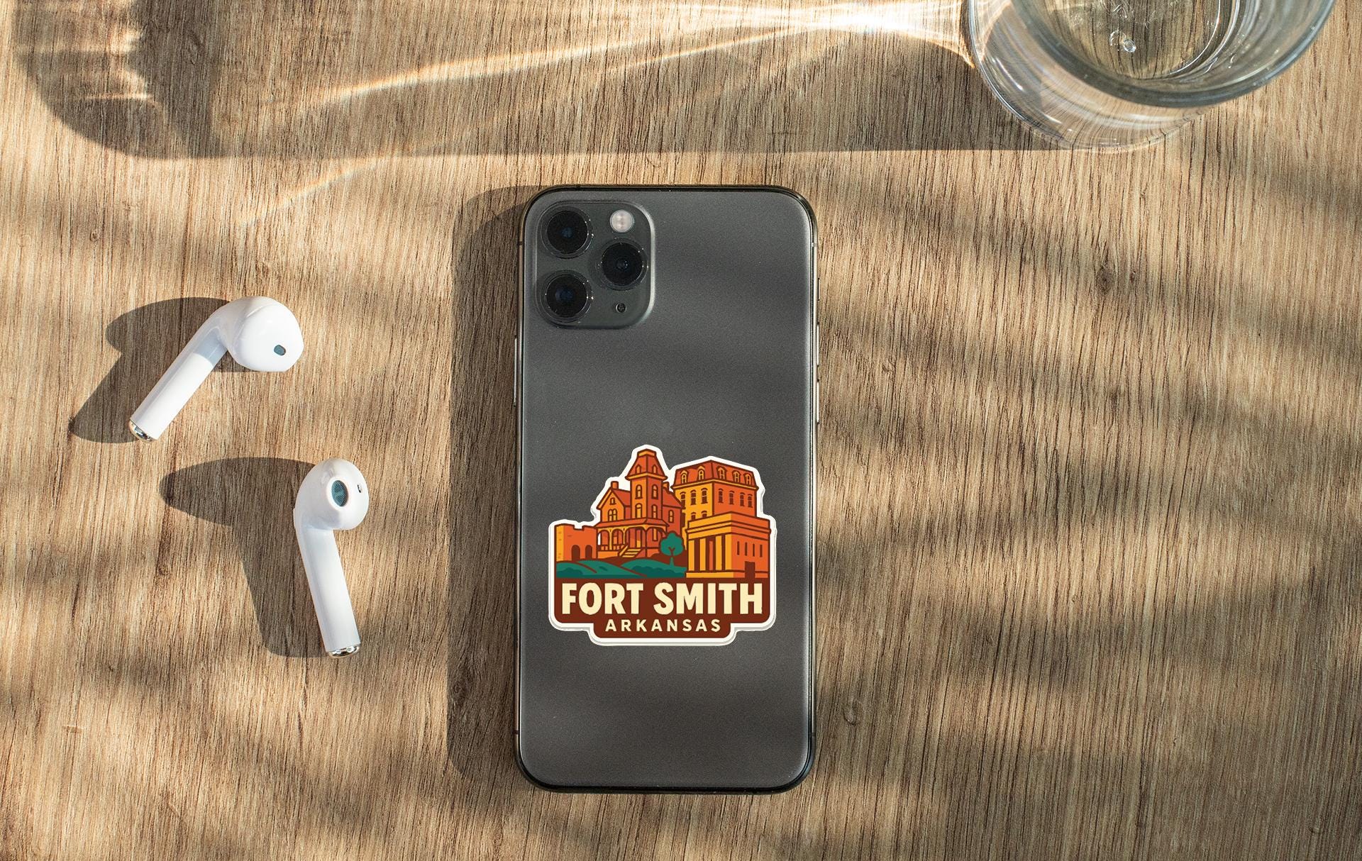 a smartphone with a sticker on the back, placed on a wooden surface alongside a pair of white earbuds and a glass of water.