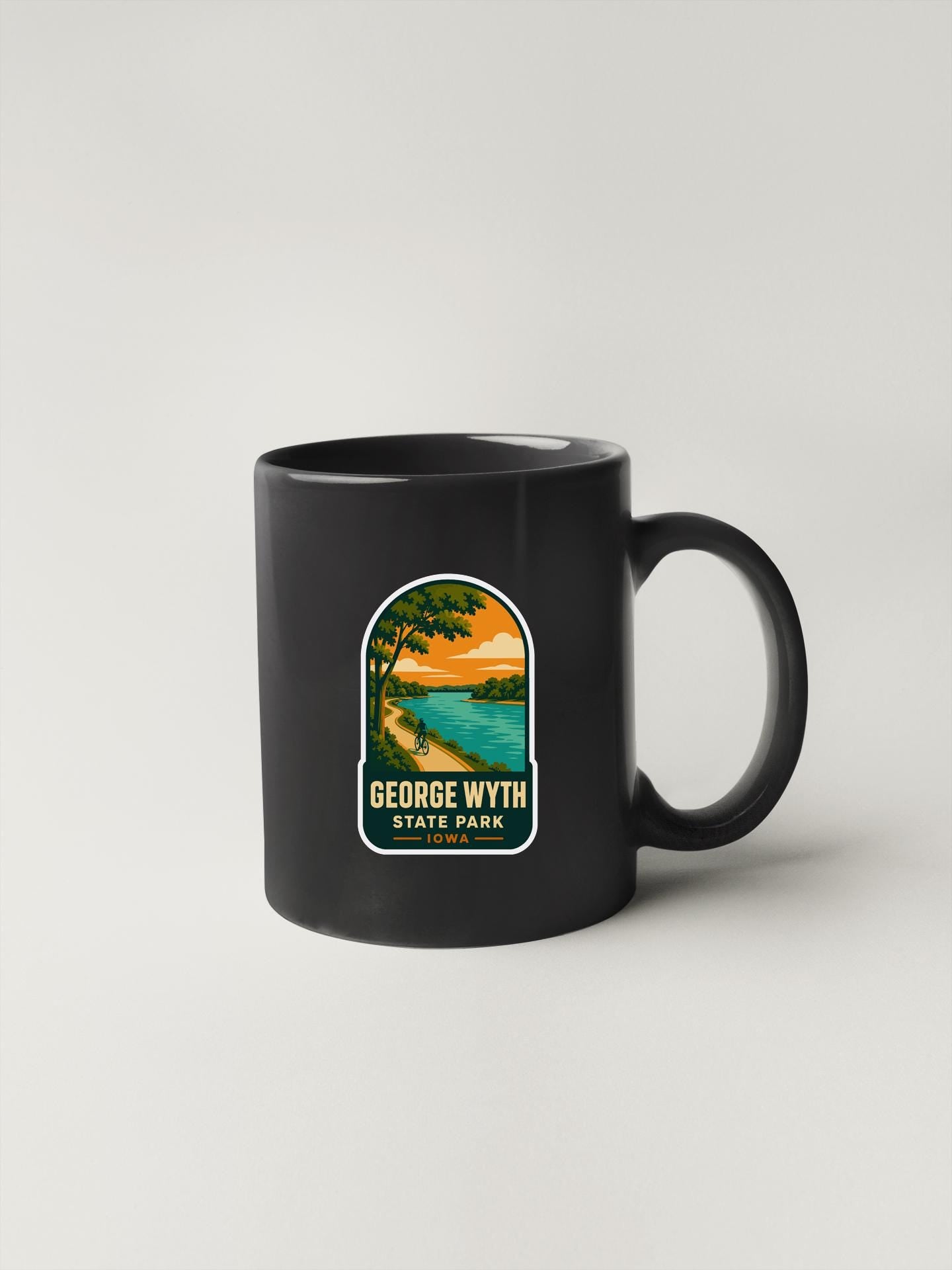 A black coffee mug with a graphic design depicting a scenic landscape, including a lake, trees, and a sunset.