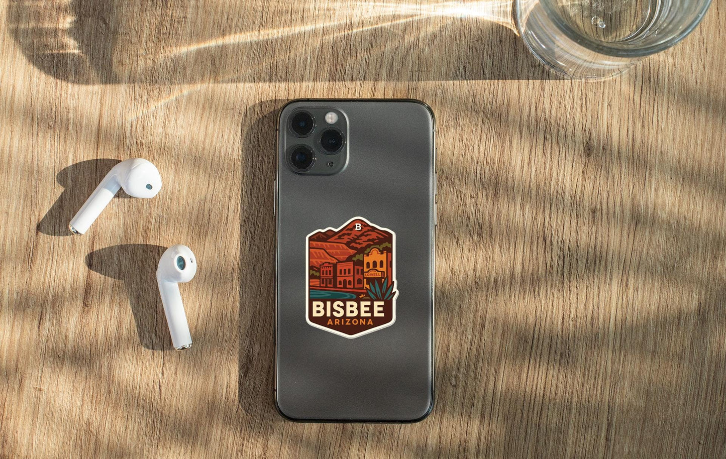 a smartphone with a sticker on the back, placed on a wooden surface alongside a pair of white earbuds and a glass of water.