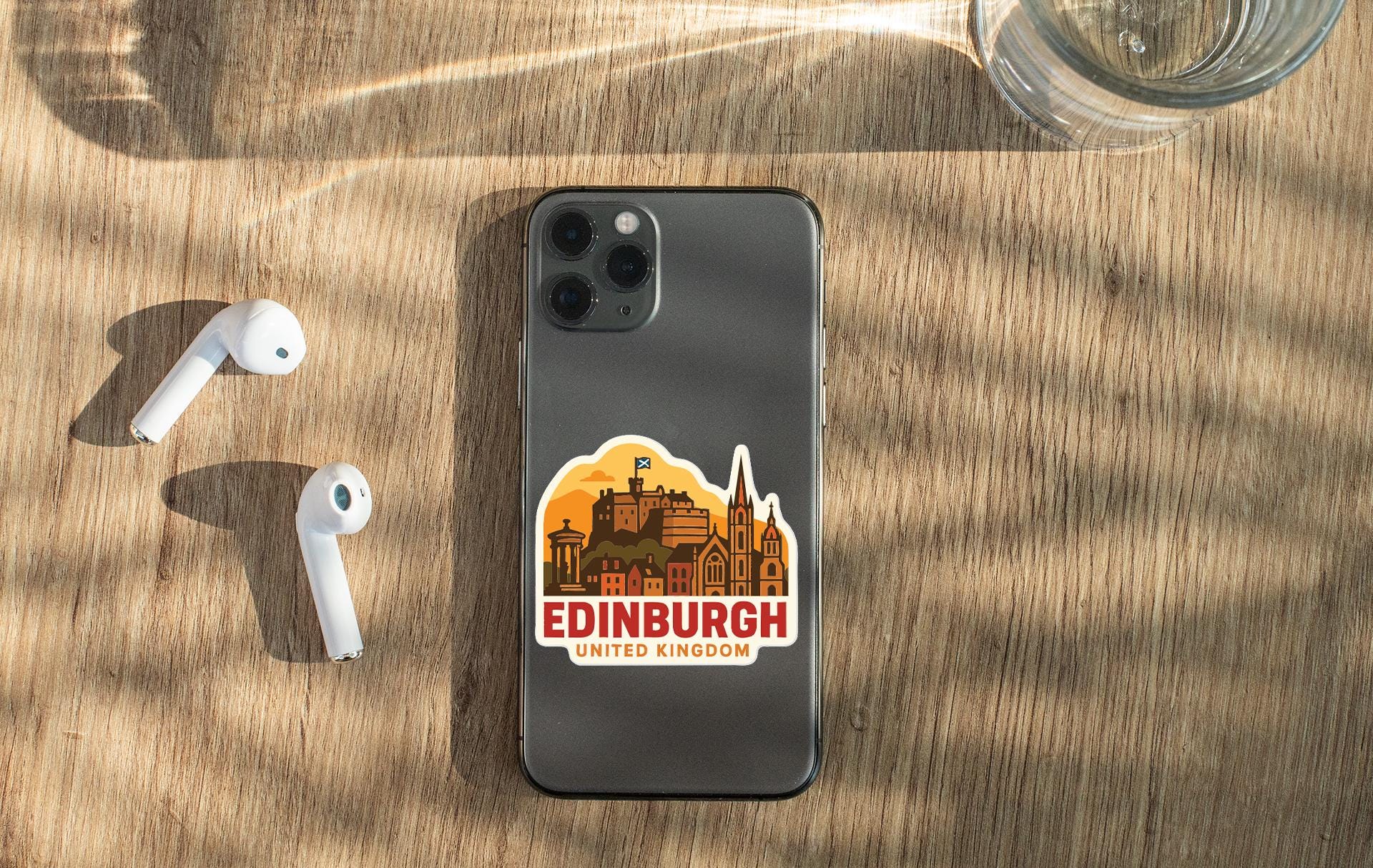 a smartphone with a sticker of Edinburgh, United Kingdom, placed on a wooden surface alongside a pair of white earbuds and a glass of water.