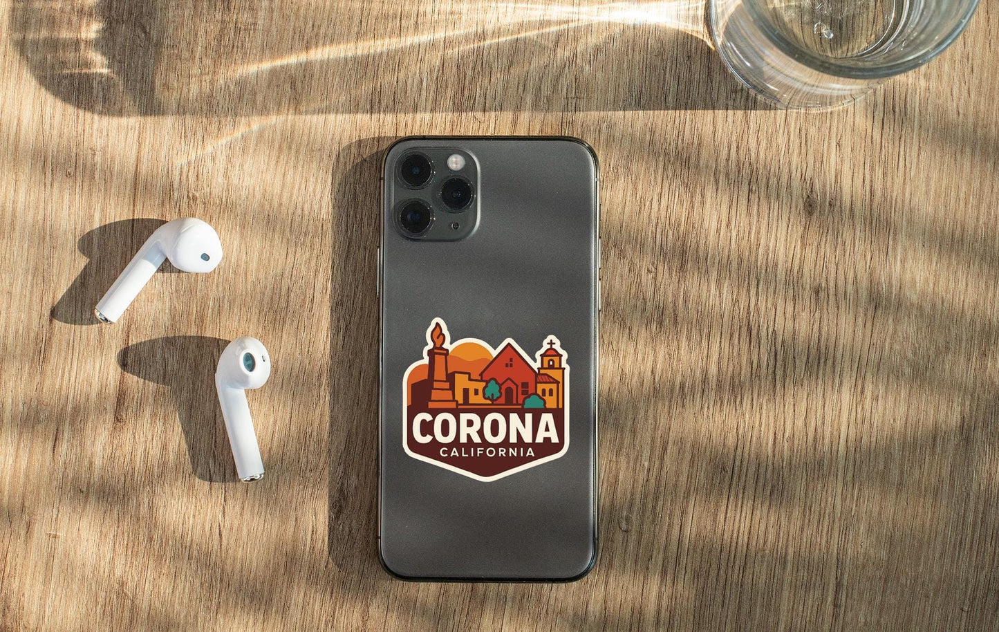 a smartphone with a sticker of Corona, California, on its back, along with a pair of white earbuds and a glass of water on a wooden surface.