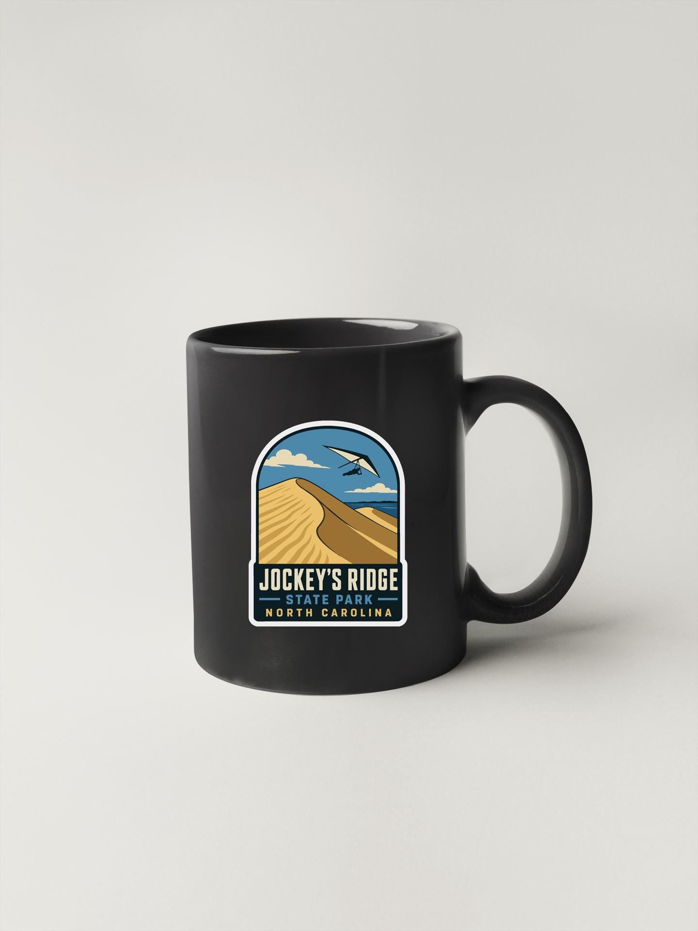 A black coffee mug with a graphic design depicting a desert landscape and the text "JOCKEY'S RIDGE" and "NORTH CAROLINA" printed on it.