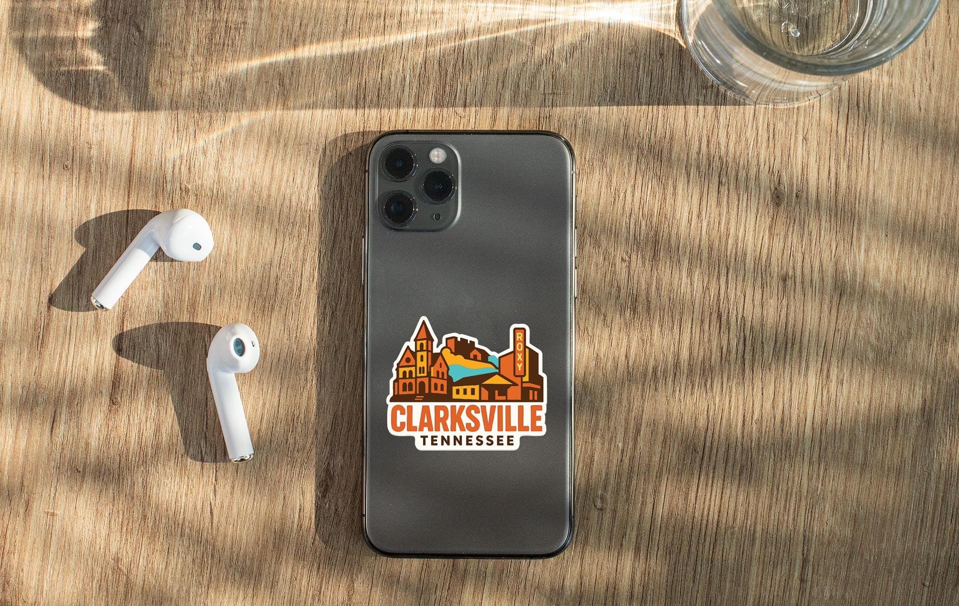 a smartphone with a sticker on it, placed on a wooden surface alongside a pair of white earbuds and a glass of water.