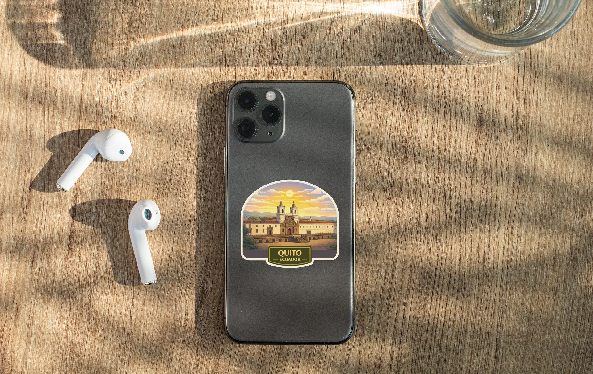 a smartphone with a sticker of Quito, the capital city of Ecuador, placed on a wooden surface alongside a pair of white earbuds and a glass of water.