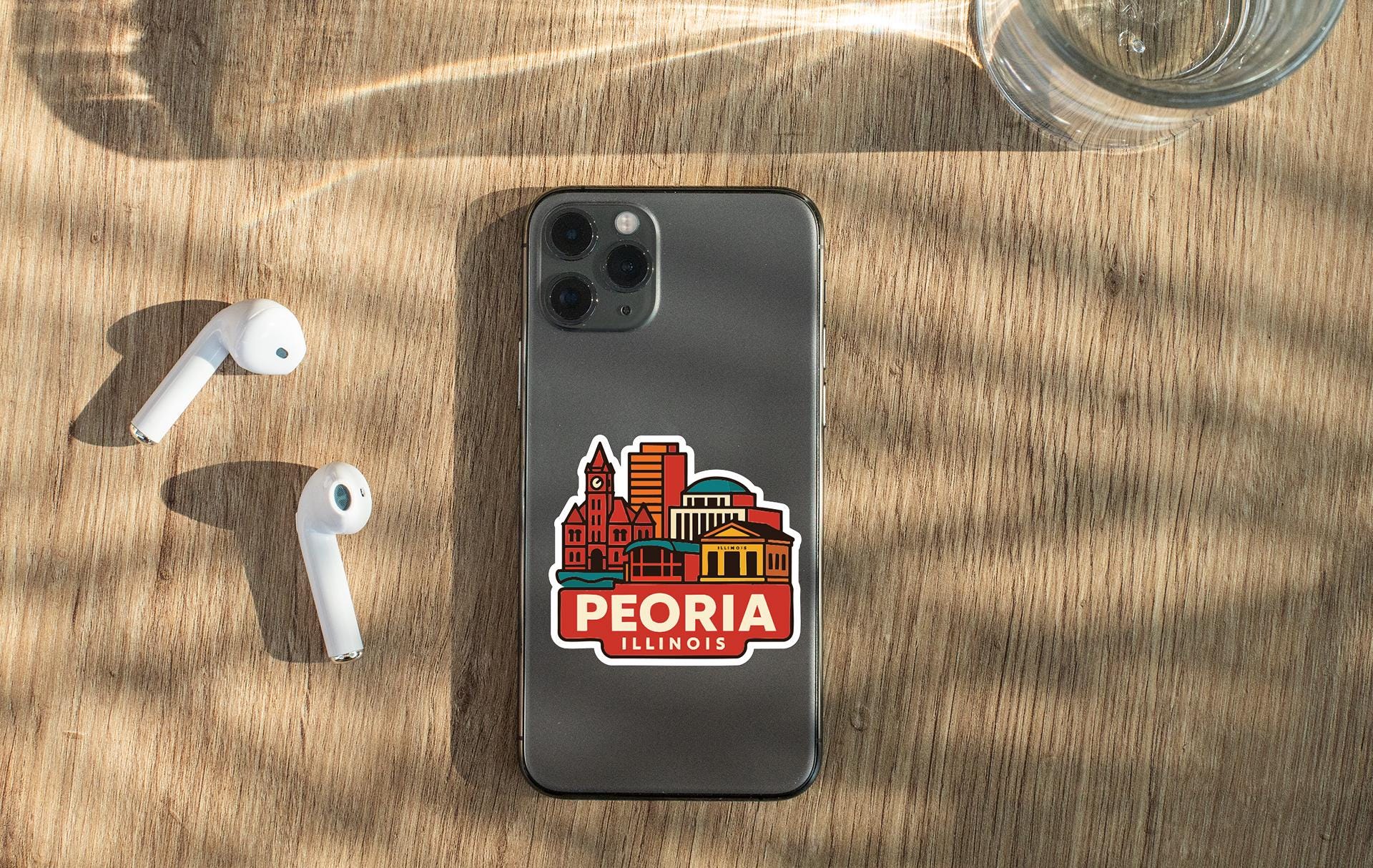a smartphone with a sticker of the city of Peoria, Illinois, placed on a wooden surface alongside a pair of white earbuds and a glass of water.