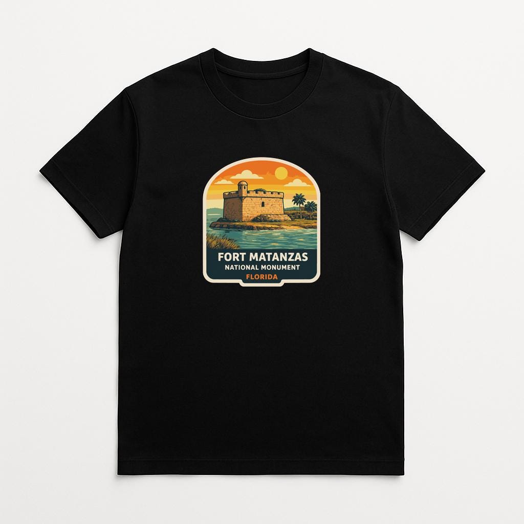 A black t-shirt with a graphic design depicting a fort or castle surrounded by palm trees and a sunset landscape.