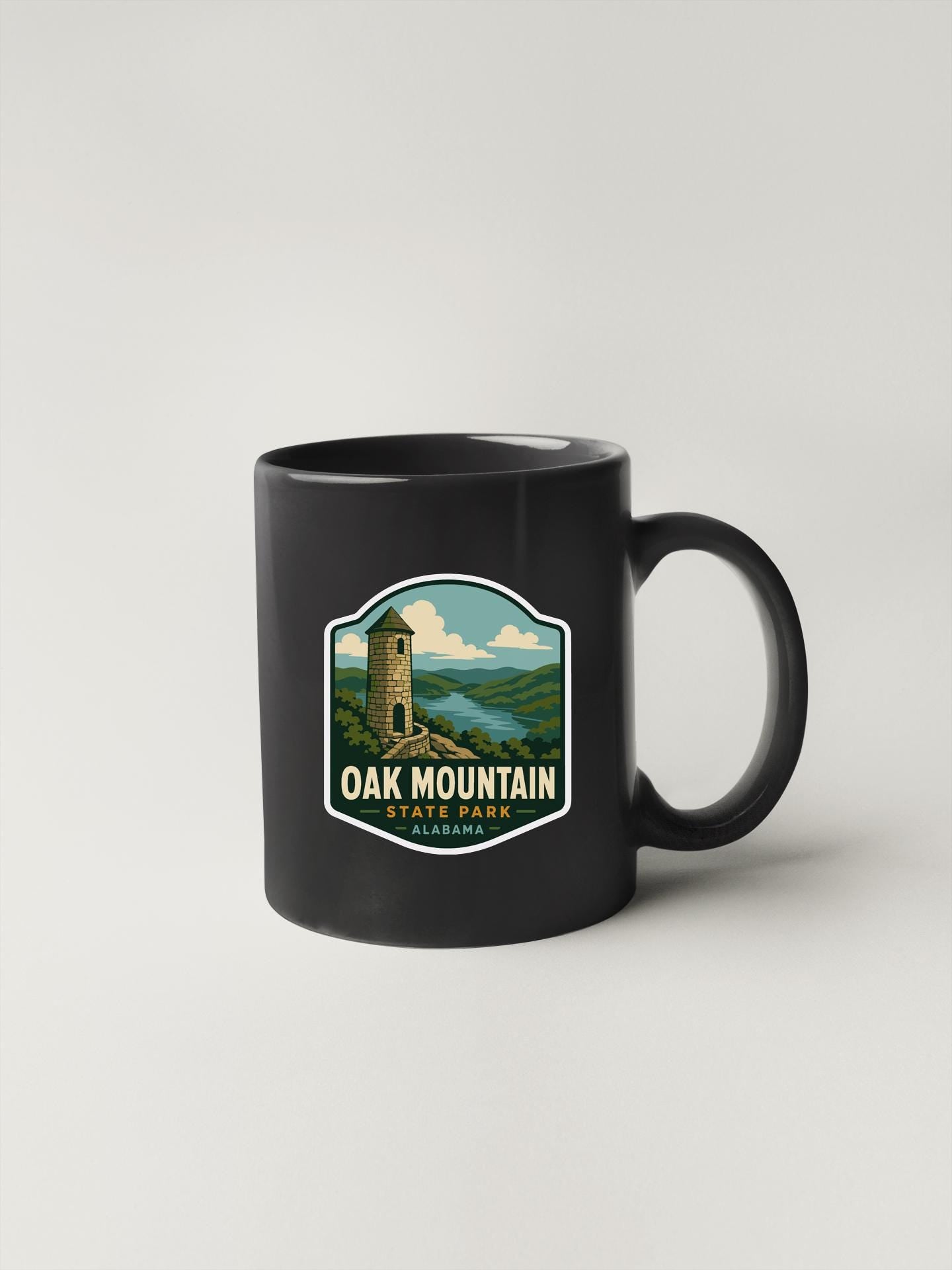 A black coffee mug with a label that says "OAK MOUNTAIN STATE PARK" and features an illustration of a tower in a scenic landscape.
