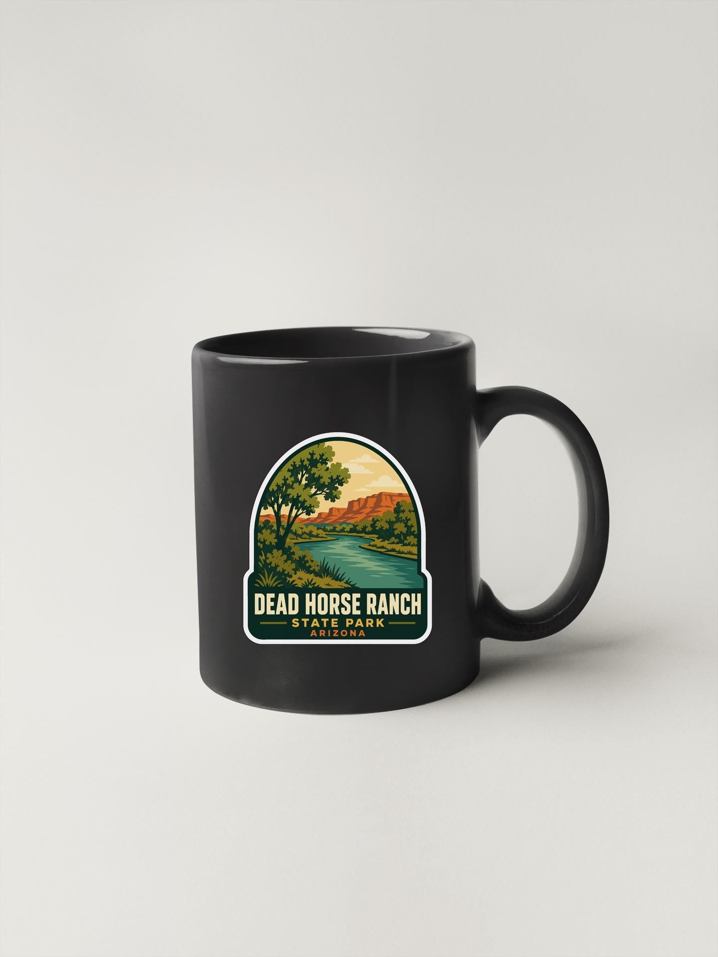 A black coffee mug with a graphic design depicting a scenic landscape, including a river, mountains, and trees.
