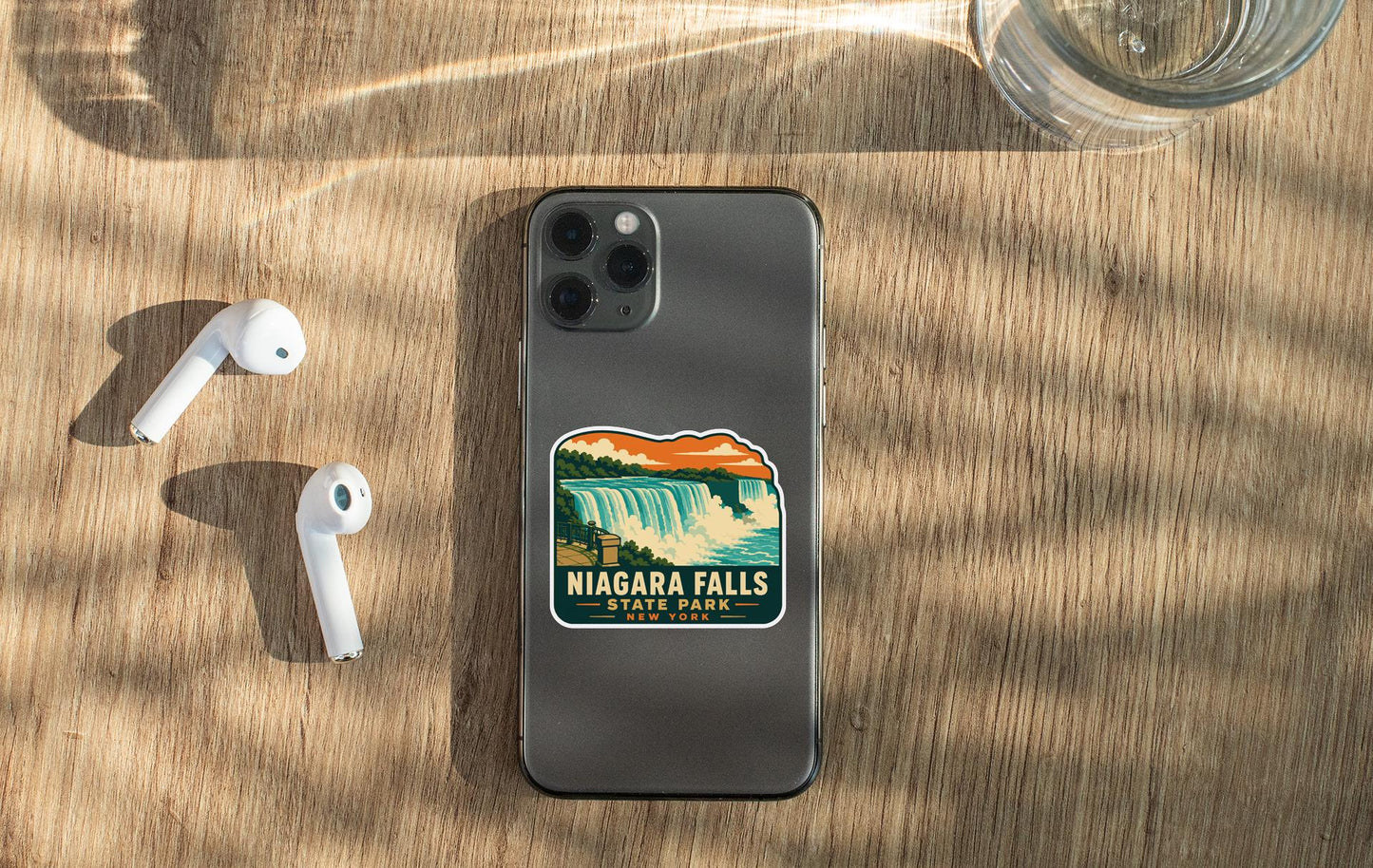 a smartphone with a sticker of Niagara Falls on it, along with a pair of white earbuds and a glass of water on a wooden surface.