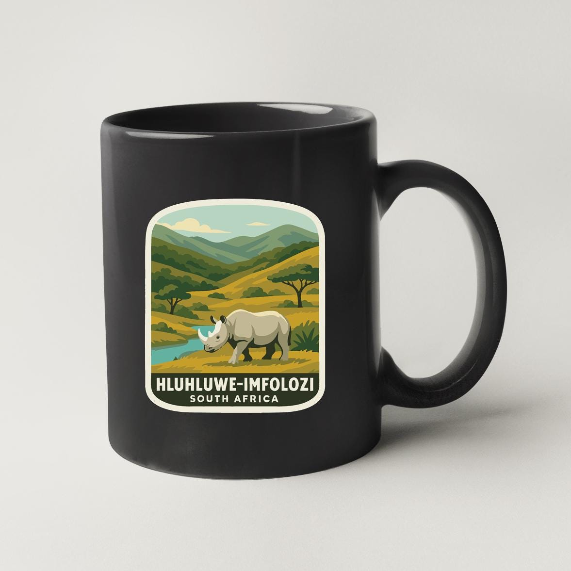 A black coffee mug with a graphic design depicting a rhinoceros in a savanna landscape, along with text that reads "HLUHUWE-IMFOLOZI SOUTH AFRICA".