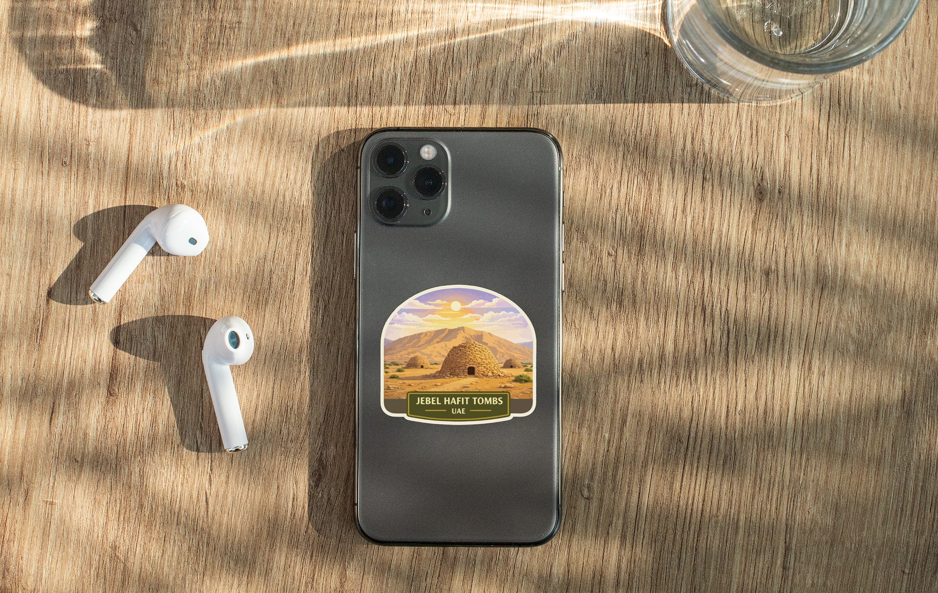 a smartphone with a sticker of a desert landscape on it, placed on a wooden surface alongside a pair of white earbuds and a glass of water.