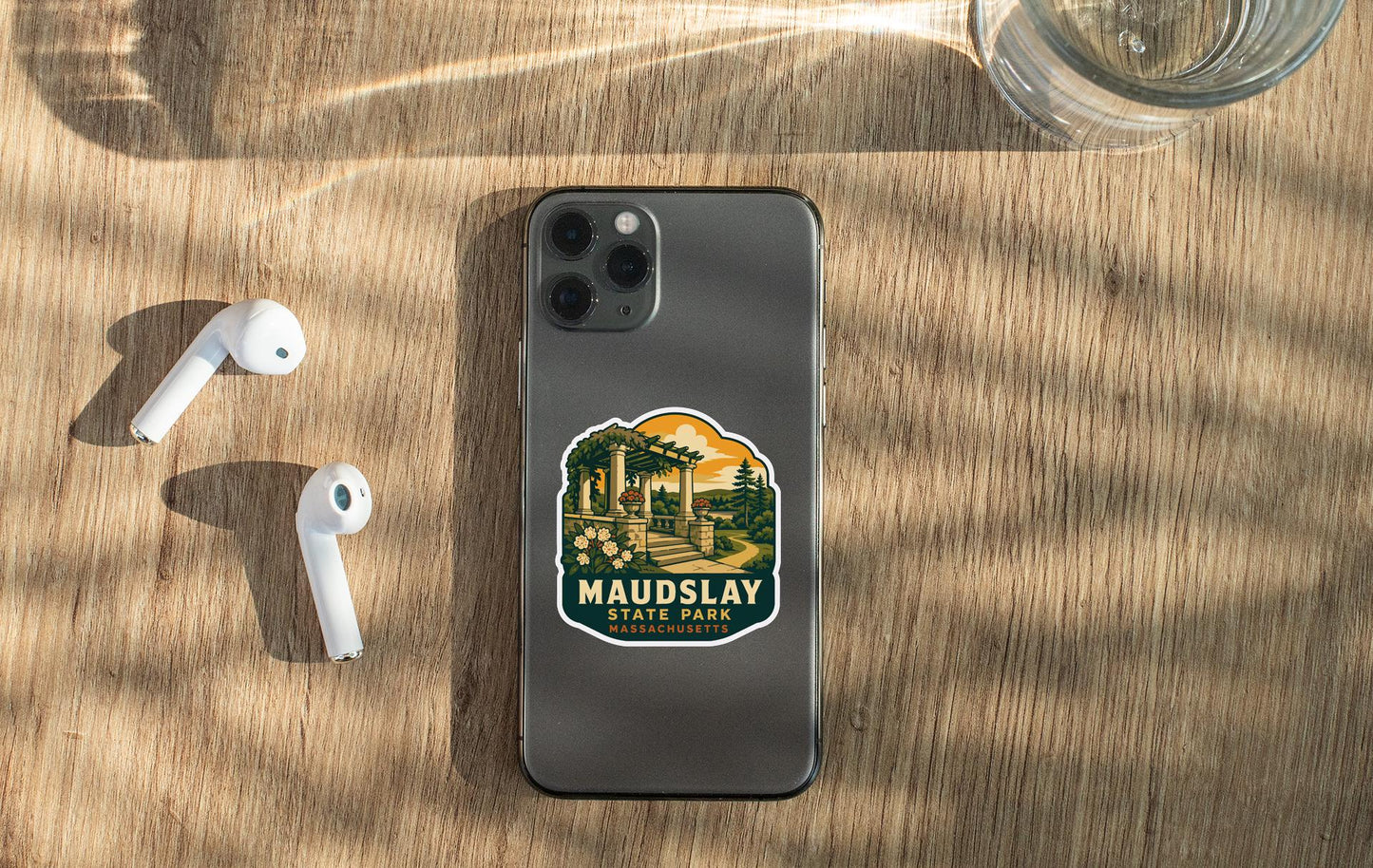 a smartphone with a sticker on it, placed on a wooden surface alongside a pair of white earbuds and a glass of water.