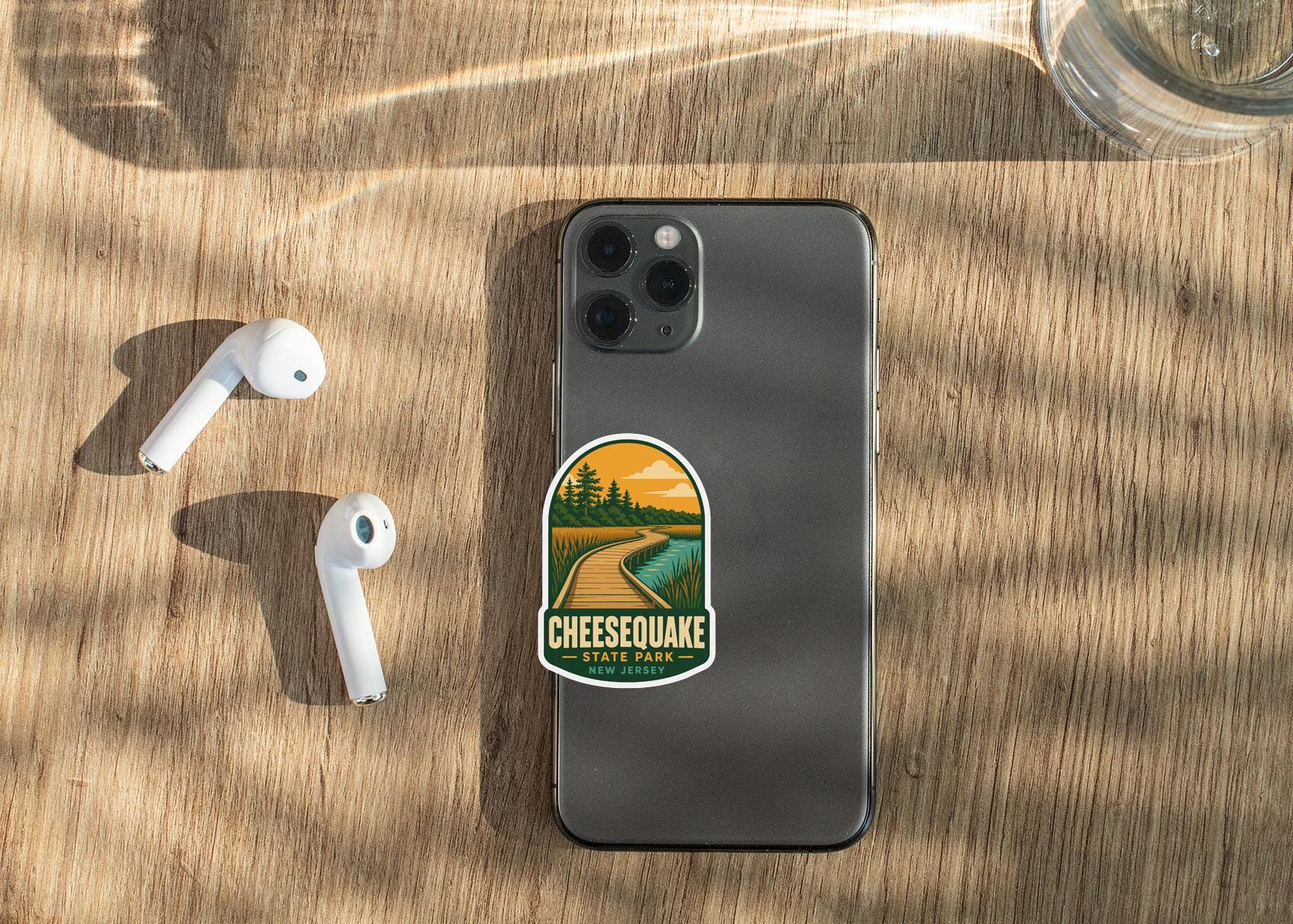 a smartphone with a sticker on it, placed on a wooden surface alongside a pair of white earbuds and a glass of water.
