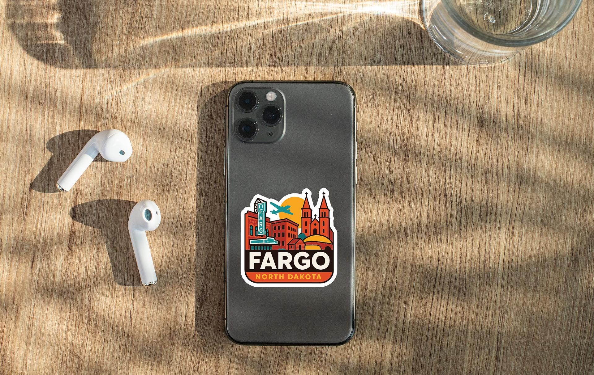 a smartphone with a sticker of a city skyline on the back, along with a pair of white earbuds and a glass of water on a wooden surface.