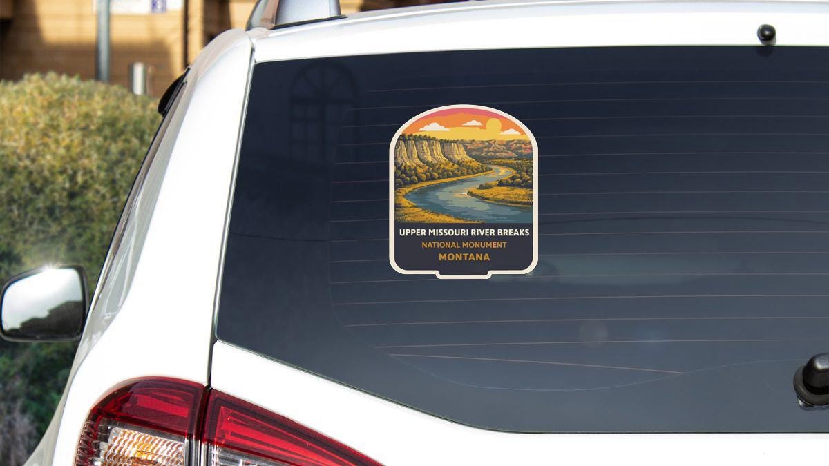 A sticker on the rear window of a white car, depicting a scenic view of the Upper Missouri River Breaks in Montana.