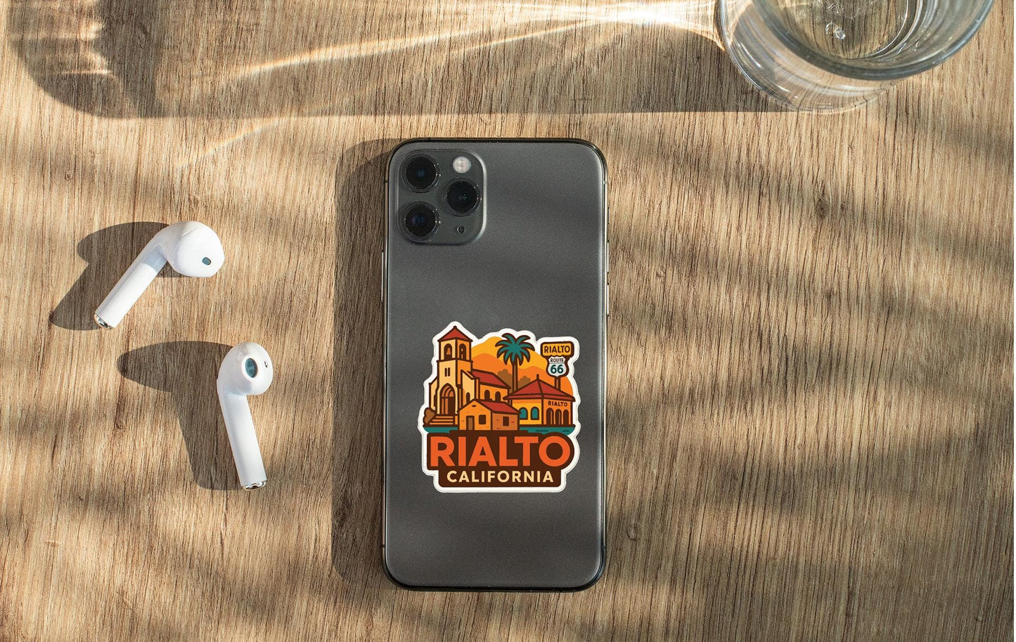 a smartphone with a sticker on the back, placed on a wooden surface alongside a pair of white earbuds and a glass of water.