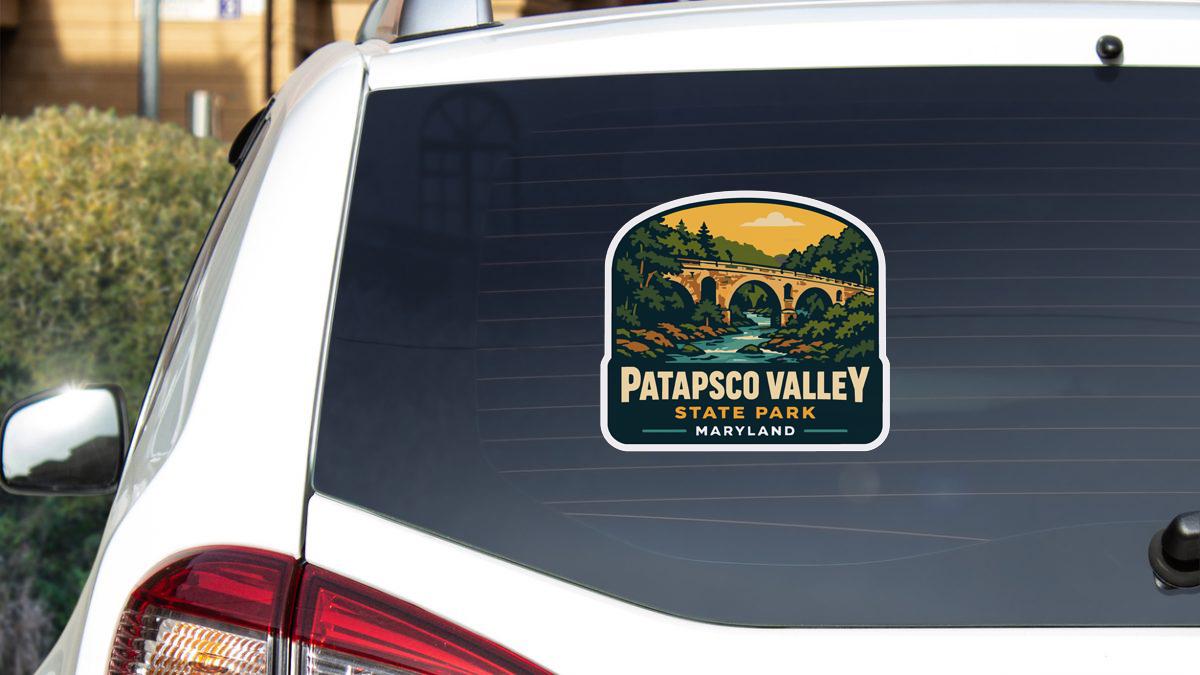 A sticker on the rear window of a white car, depicting a scenic landscape with a bridge and trees, indicating that it is a sticker for Patapsco Valley State Park in Maryland.