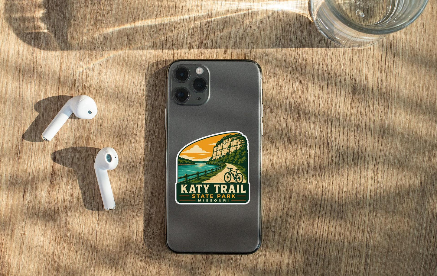 a smartphone with a sticker on the back, placed on a wooden surface alongside a pair of white earbuds and a glass of water.