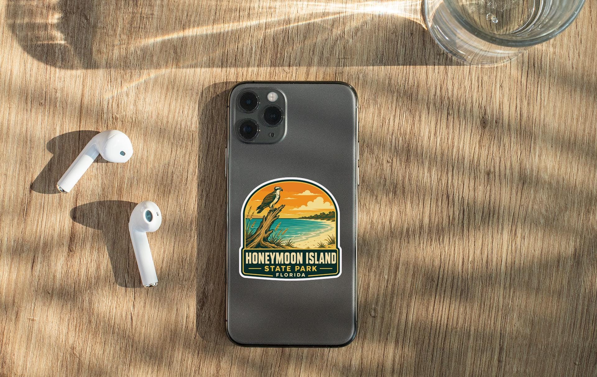 a smartphone with a sticker on it, placed on a wooden surface alongside a pair of white earbuds and a glass of water.