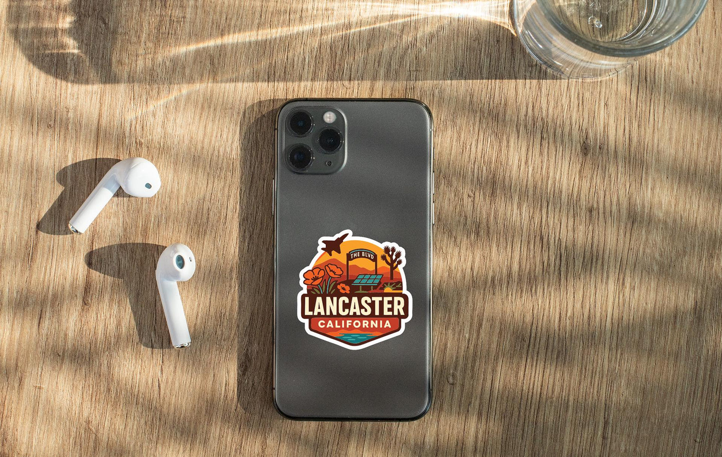 a smartphone with a sticker on the back, placed on a wooden surface alongside a pair of white earbuds and a glass of water.