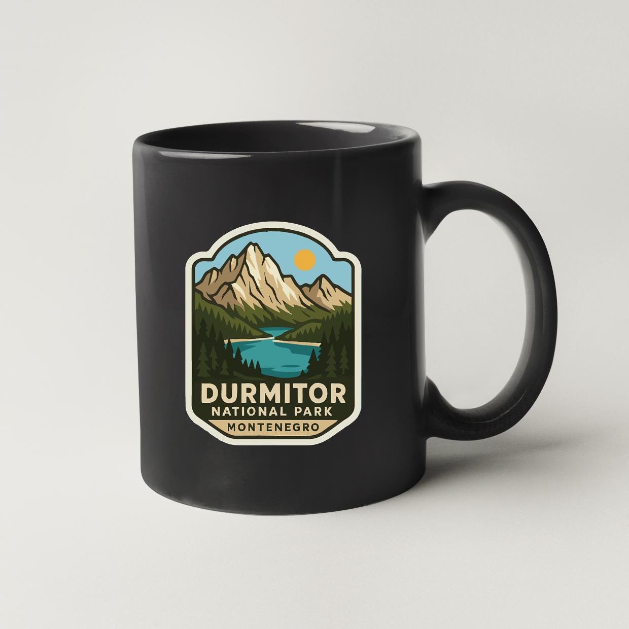 A black coffee mug with a graphic design depicting a mountain landscape, a lake, and a sun. The text on the mug reads "DURMITOR NATIONAL PARK MONTENEGRO".