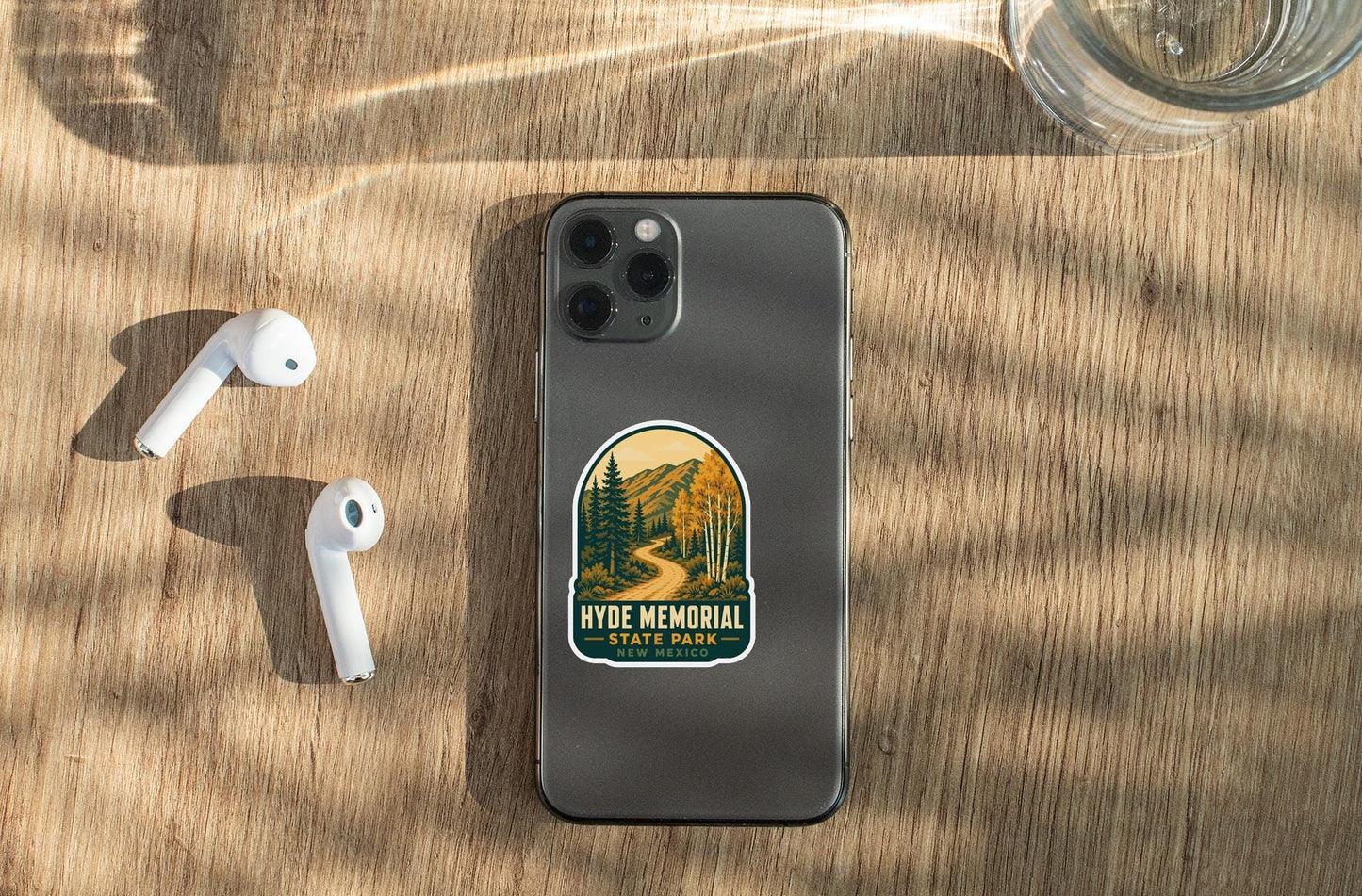 a smartphone with a sticker on it, placed on a wooden surface alongside a pair of white earbuds and a glass of water.