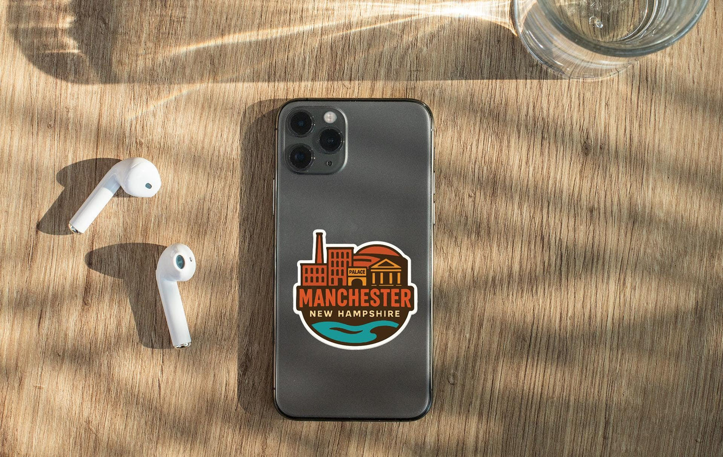 a smartphone with a Manchester New Hampshire sticker on it, placed on a wooden surface alongside a pair of white earbuds and a glass of water.