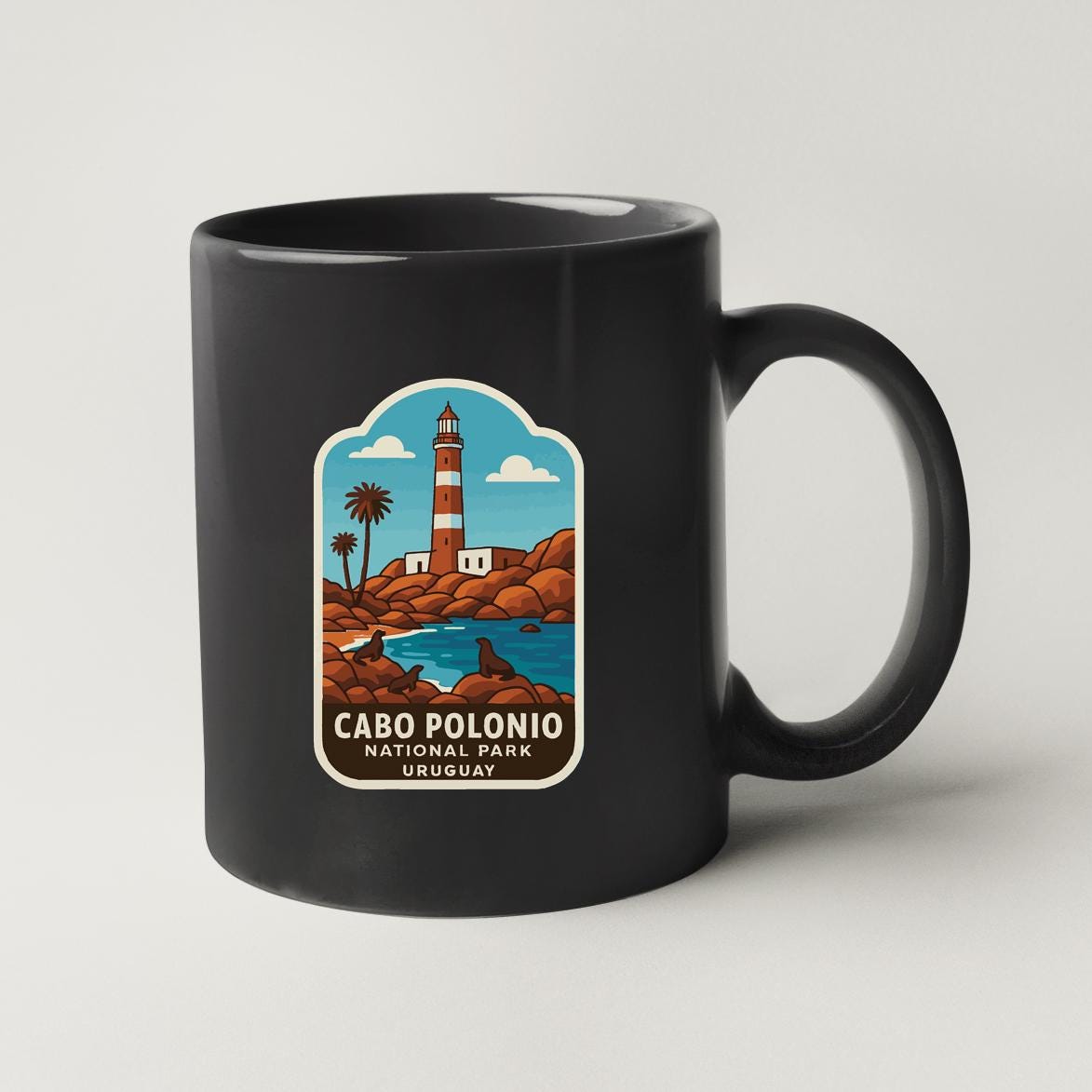 A black coffee mug with a label that features an illustration of a lighthouse and a beach scene, along with the text "CABO POLONIO NATIONAL PARK UOGUAY".