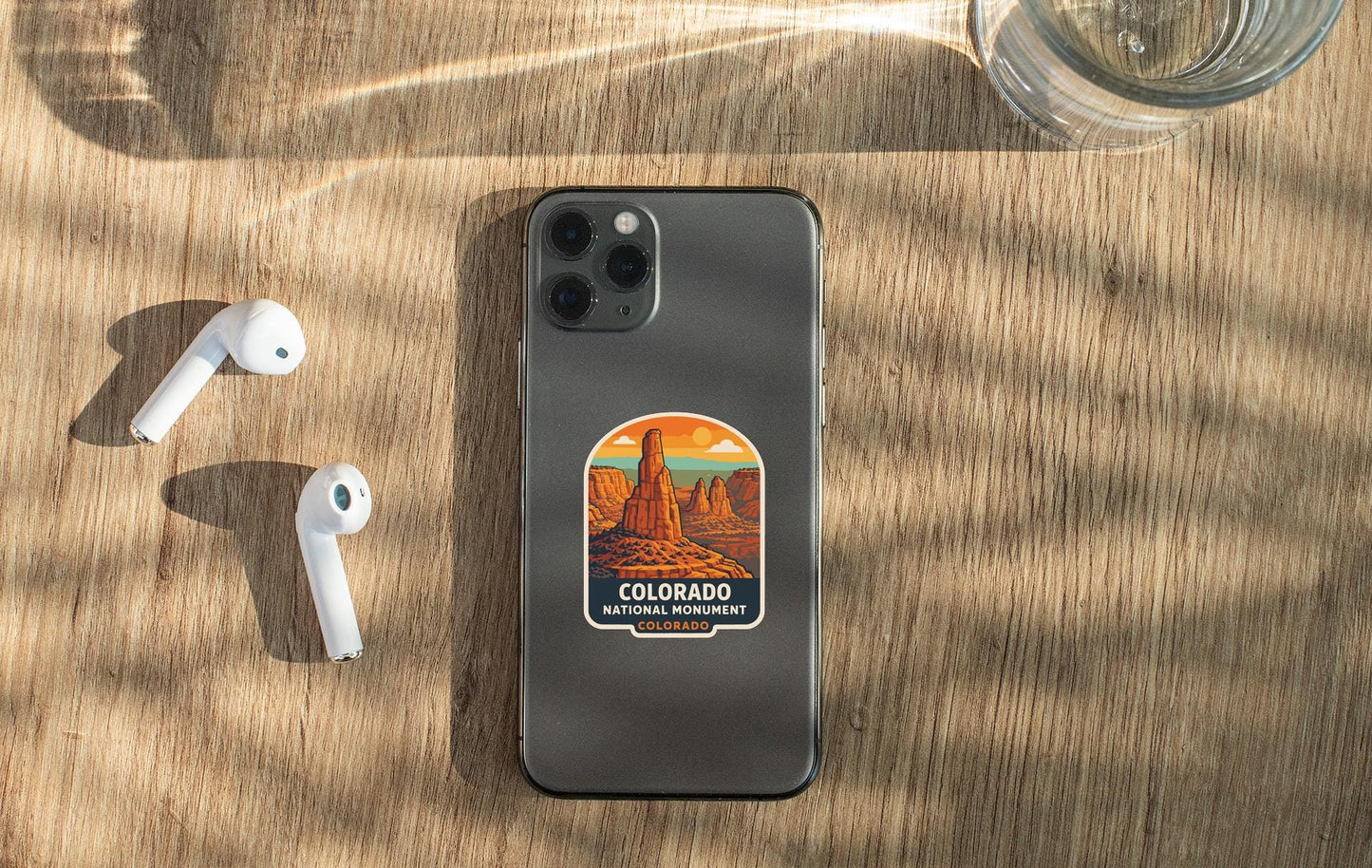 a smartphone with a Colorado National Forest sticker on its back, placed on a wooden surface alongside a pair of white earbuds and a glass of water.