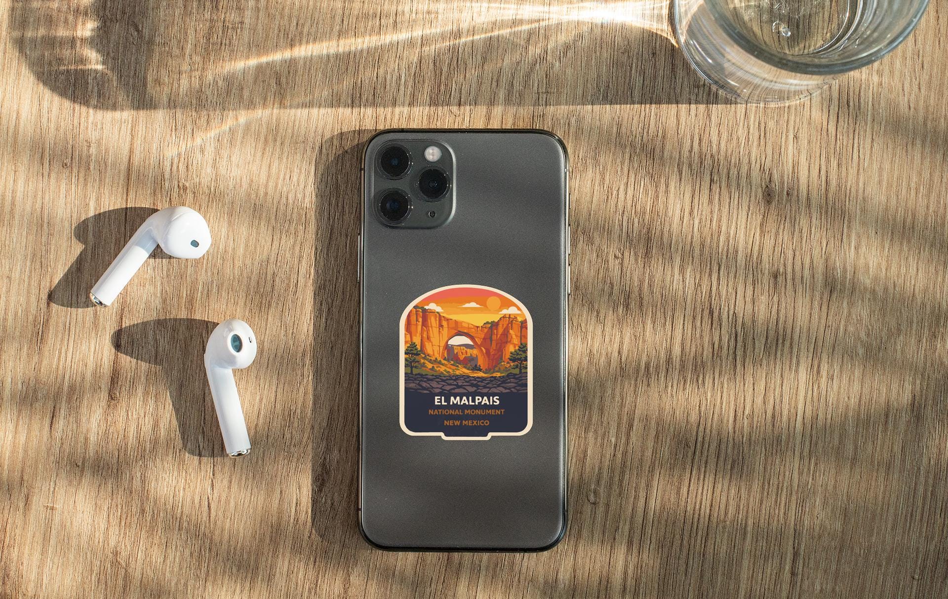 a smartphone with a sticker of a scenic landscape, a pair of white earbuds, and a glass of water on a wooden surface.