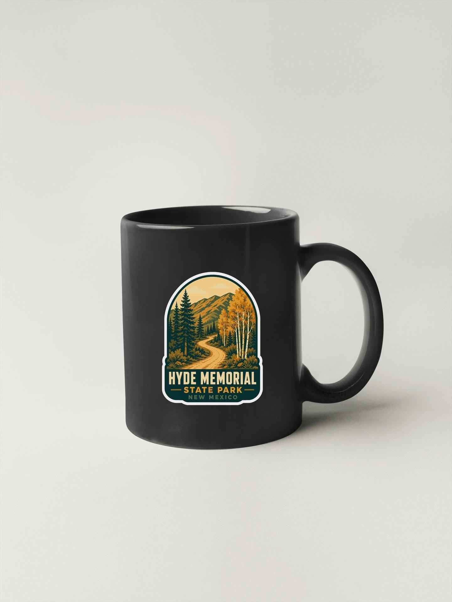 A black coffee mug with a graphic design featuring a scenic landscape with trees, mountains, and a winding road.