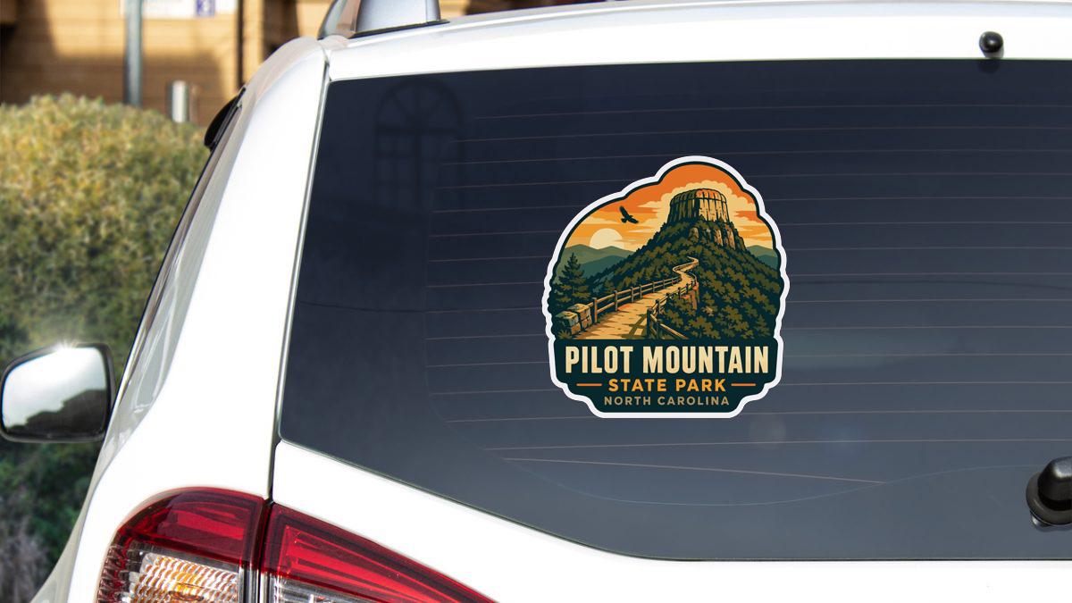 a sticker on the rear window of a white vehicle, depicting a scenic mountain landscape with a trail leading up to a tower.