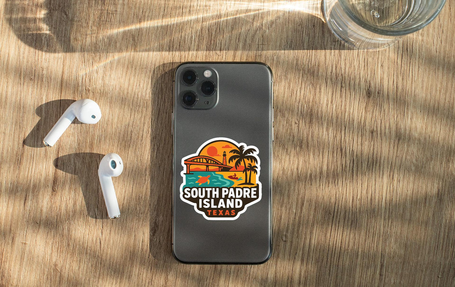 a smartphone with a sticker on it, placed on a wooden surface alongside a pair of white earbuds and a glass of water.