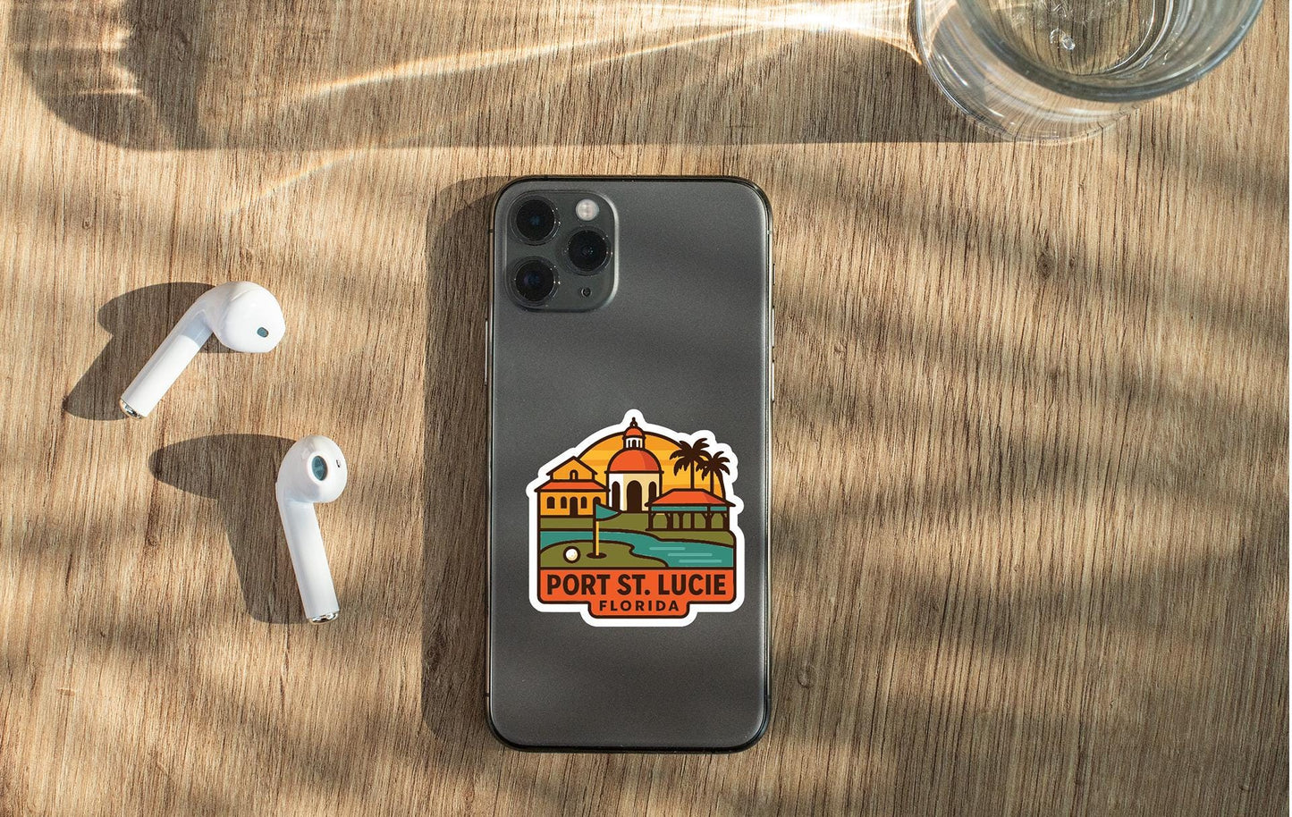 a smartphone with a sticker of Port St. Lucie, Florida, placed on a wooden surface alongside a pair of white earbuds and a glass of water.