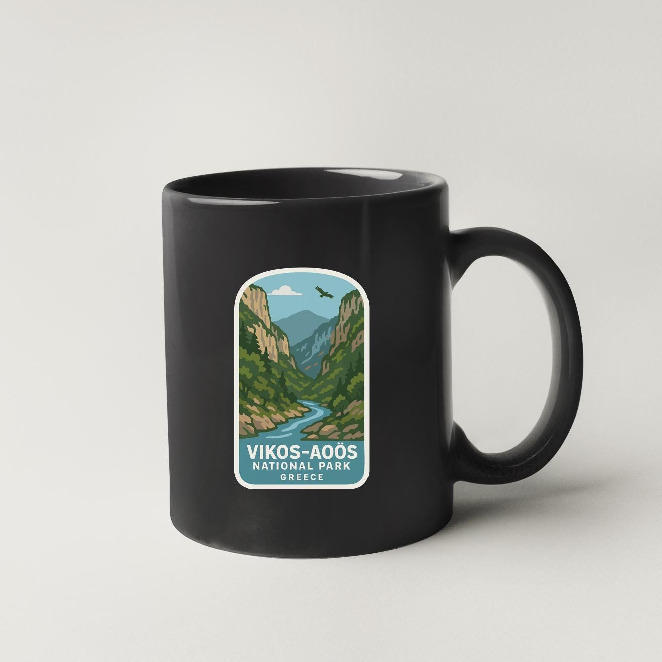 A black coffee mug with a label that features an illustration of a mountain landscape with a river, along with the text "VIKOS-AOOS NATIONAL PARK GREECE".