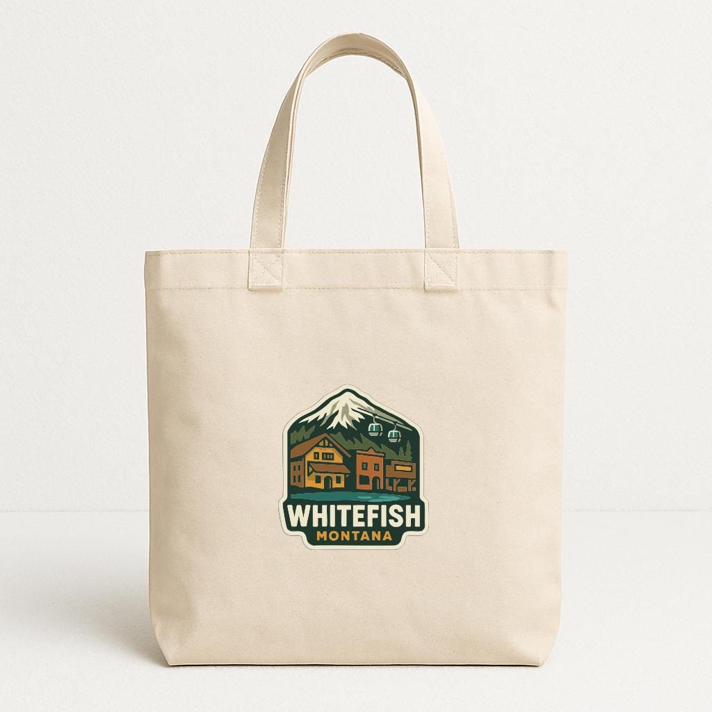 A beige canvas tote bag with a graphic design featuring a mountain, buildings, and text that says "WHITEFISH MONTANA".