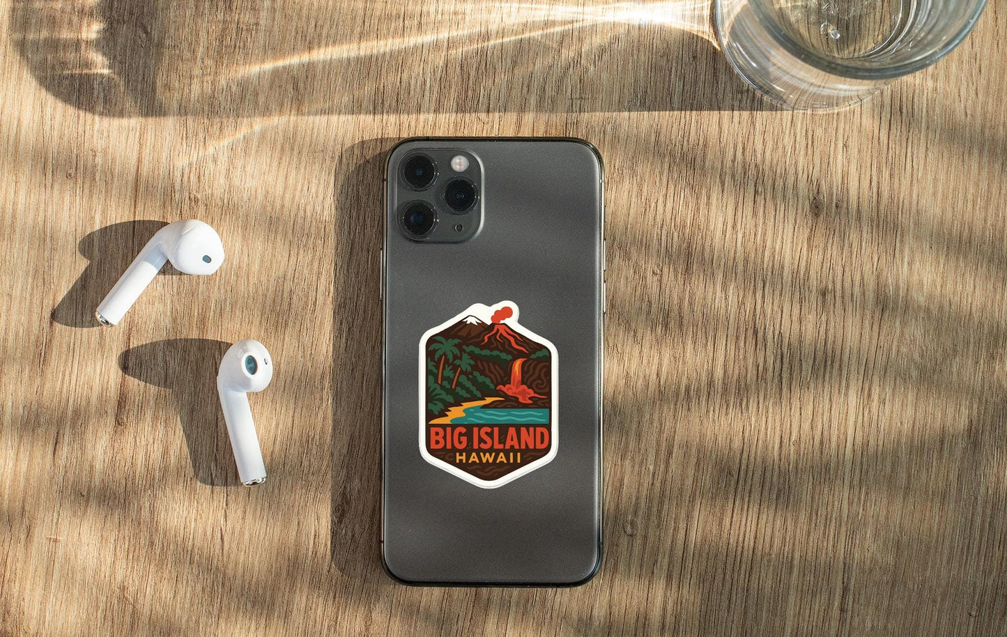 a smartphone with a sticker of Big Island, Hawaii, placed on a wooden surface alongside a pair of white earbuds and a glass of water.